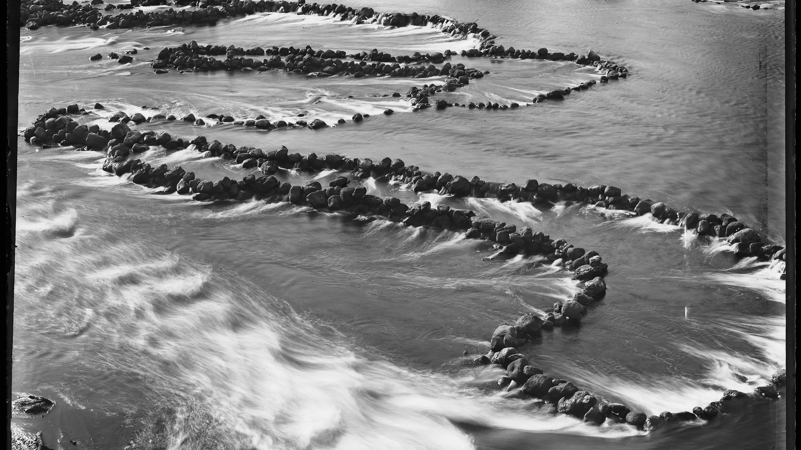 A silver gelatin dry plate glass negative in landscape format. A silver gelatin dry plate glass negative in landscape format. The image shows fish traps in a section of the Darling River. The fish traps consist of low stone walls constructed on the river bed. The walls are arranged to form cul de sacs that trap the fish. The Tyrrell Collection Inventory records the above caption but does not attribute the negative to a studio.