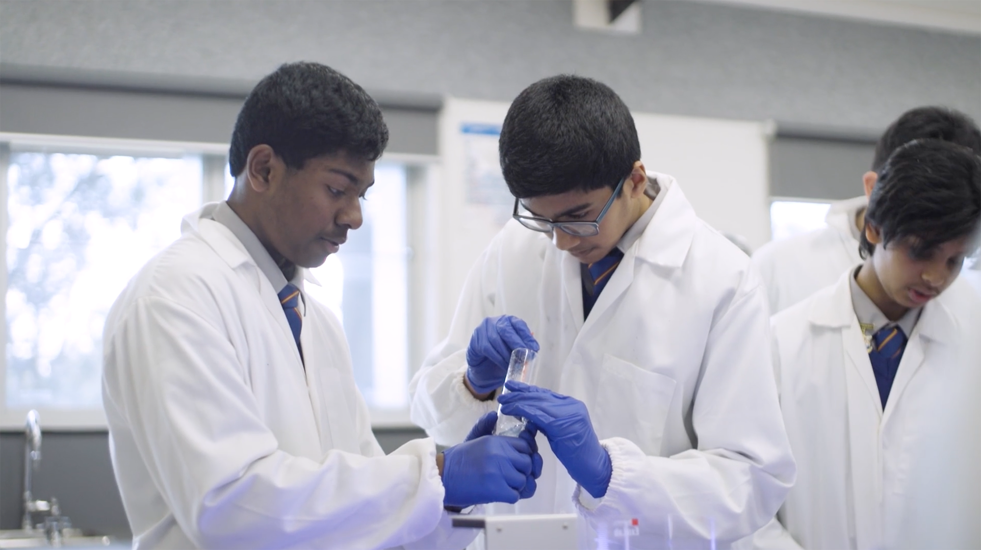 Two students wearing lab coats and blue gloves work together on an experiment.
