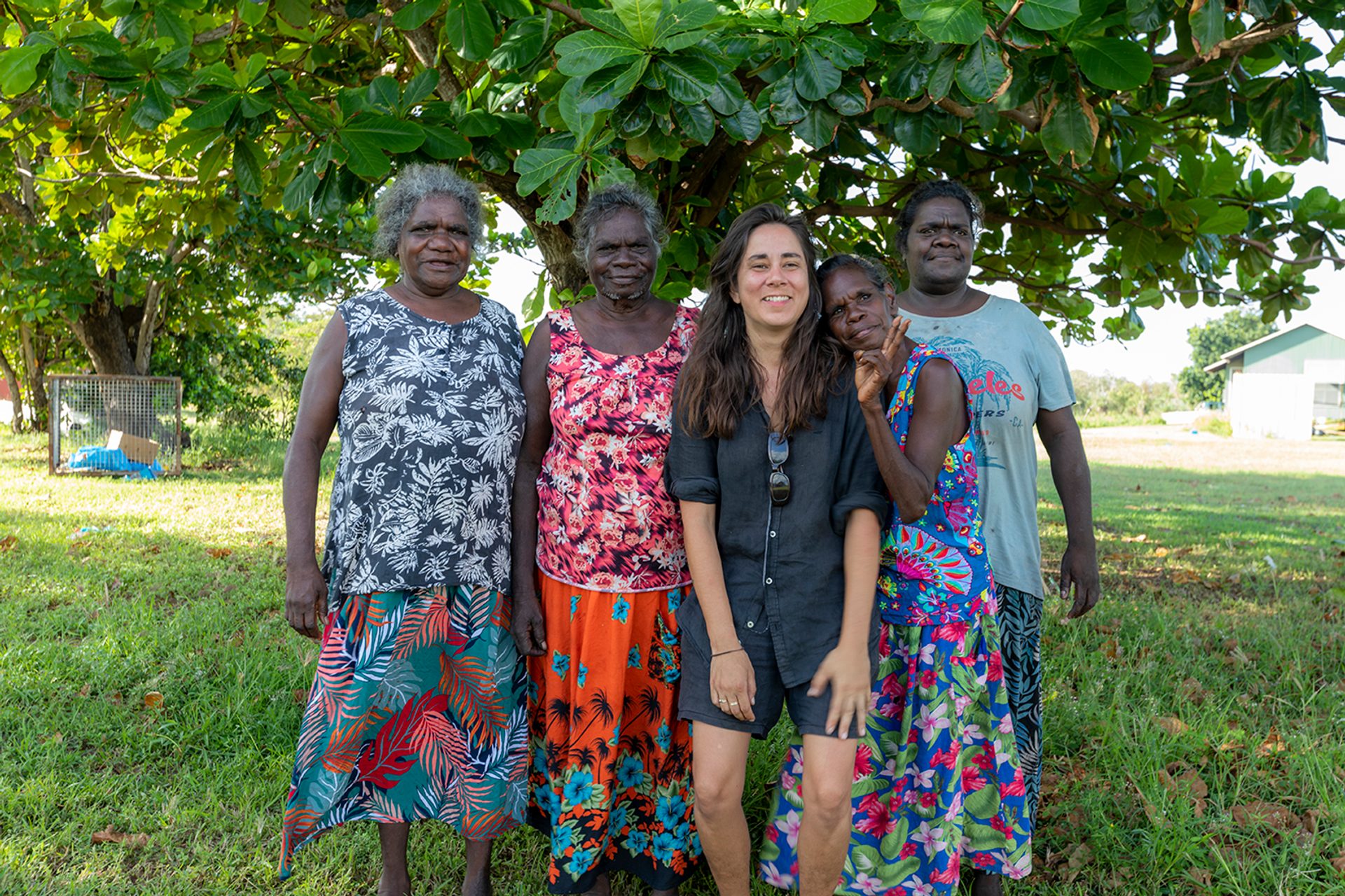 A group of five women stand outside on the grass, in front of a large, green leafy tree.