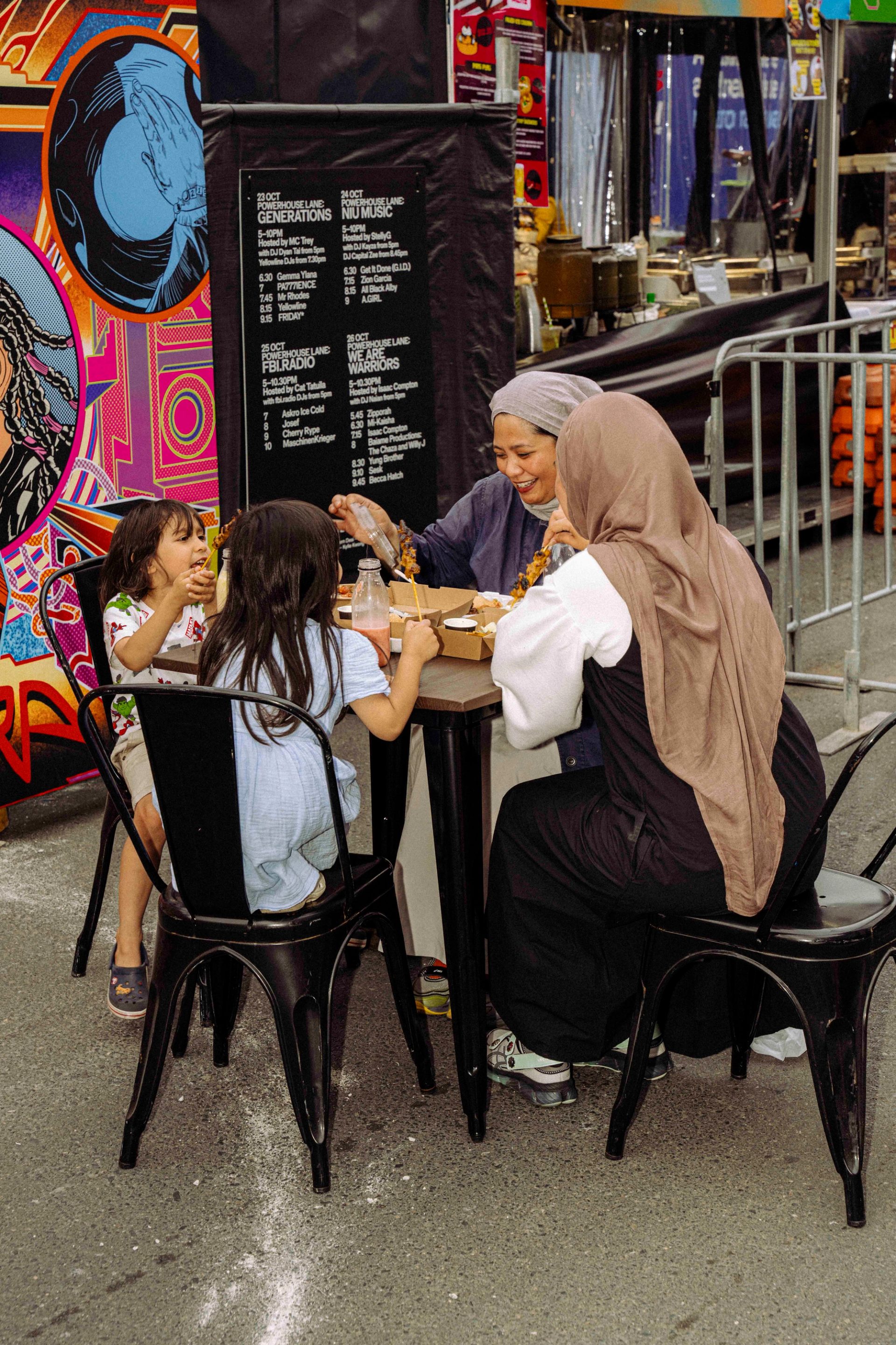 Two adults wearing veils, and two children share a meal at a small black metal table in an outdoor market, with a colourful mural in the background.