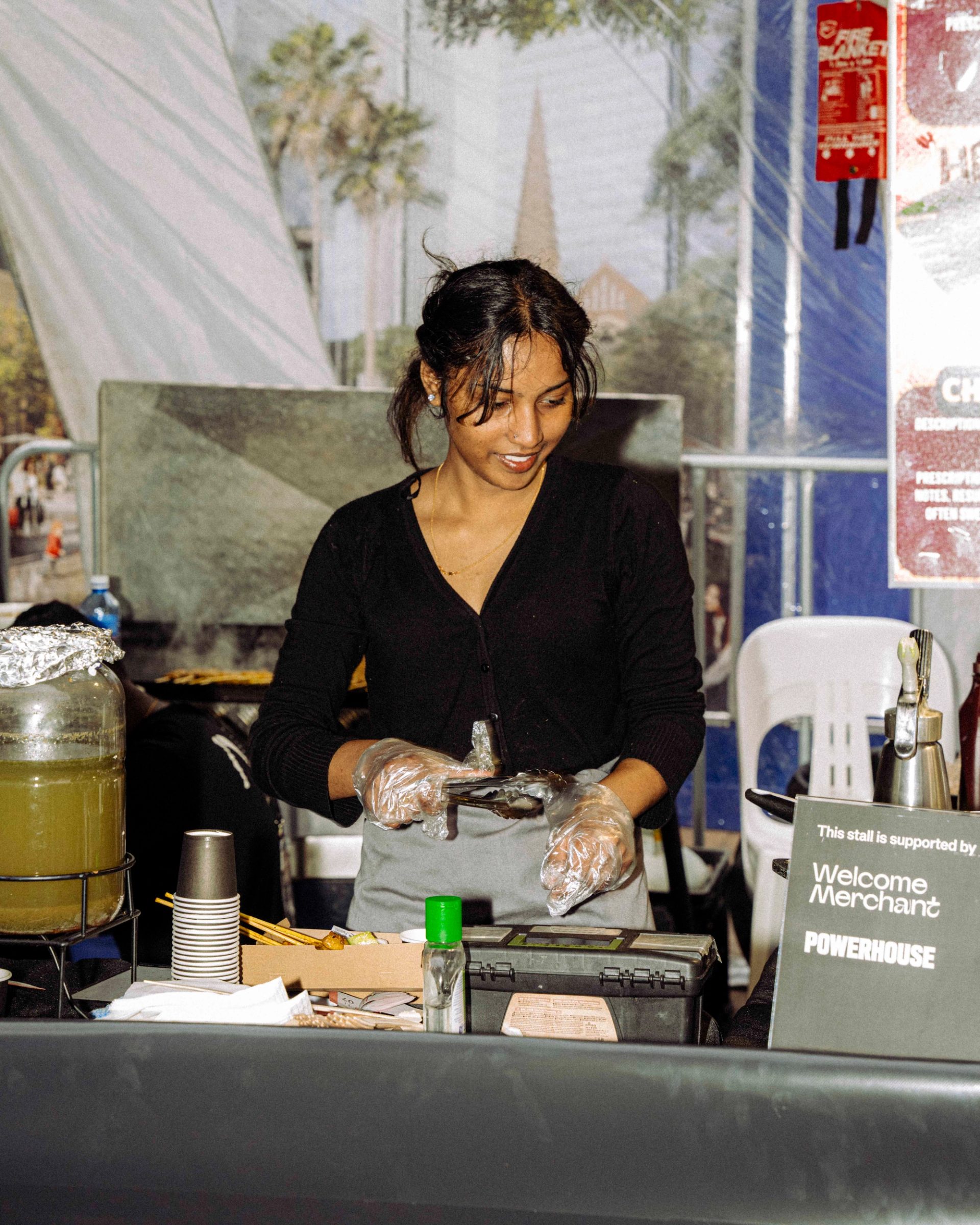 A smiling person in a black top and gloves prepares food at a stall, with kitchen items visible in the background, and a ‘Welcome Merchant’ sign on the counter.