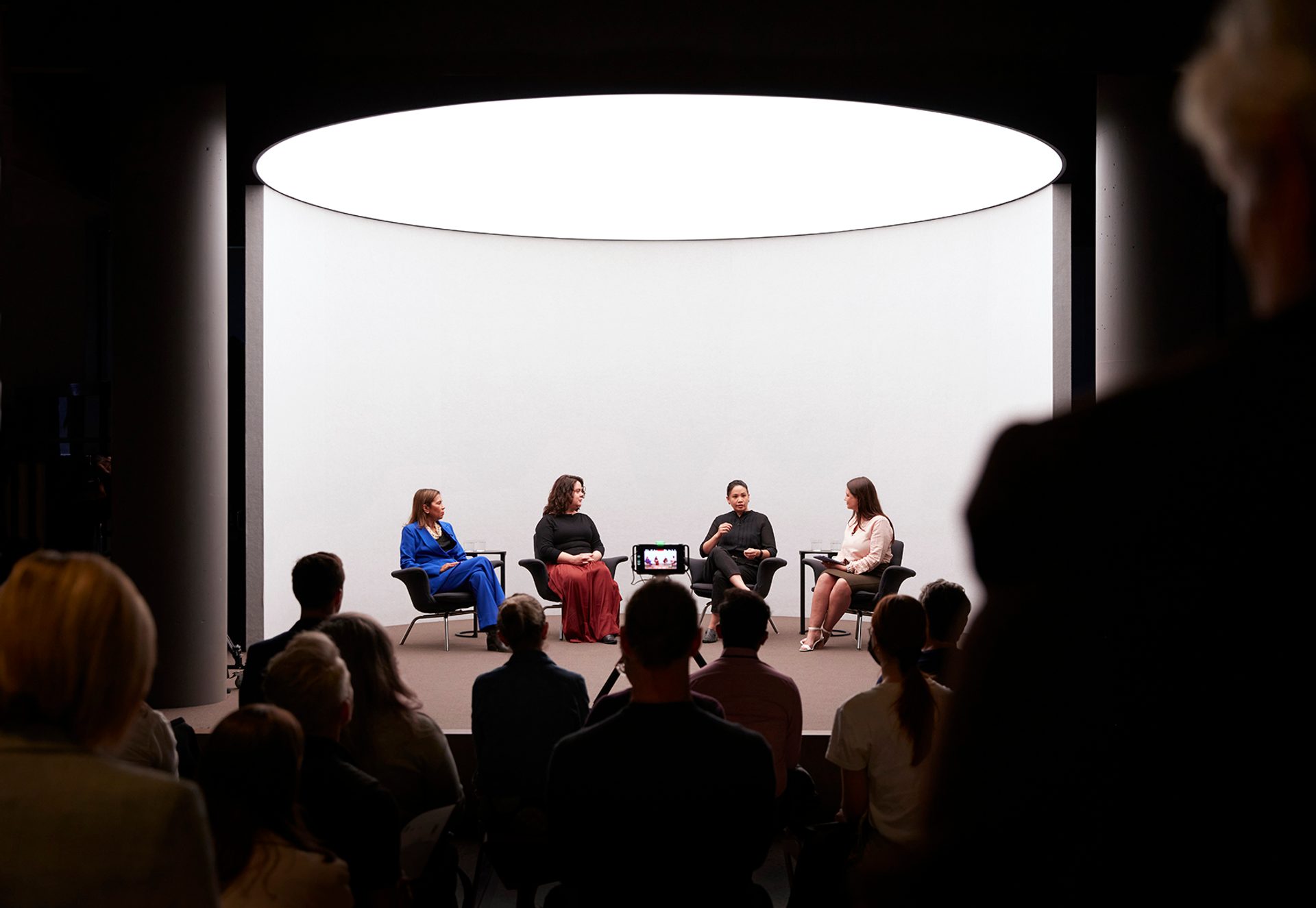 Four figures sit on a stage with a white background talking, there is a crowd watching the talk.