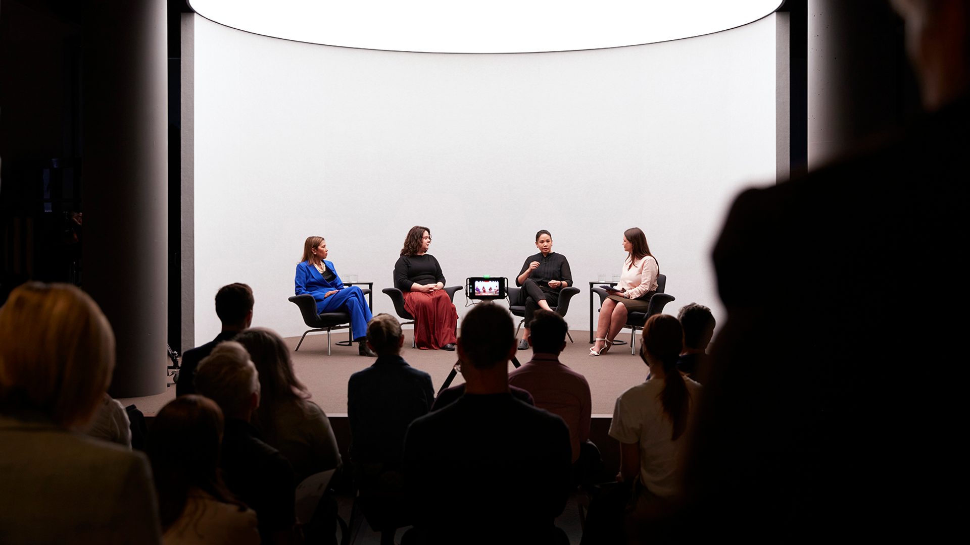 Four figures sit on a stage with a white background talking, there is a crowd watching the talk.