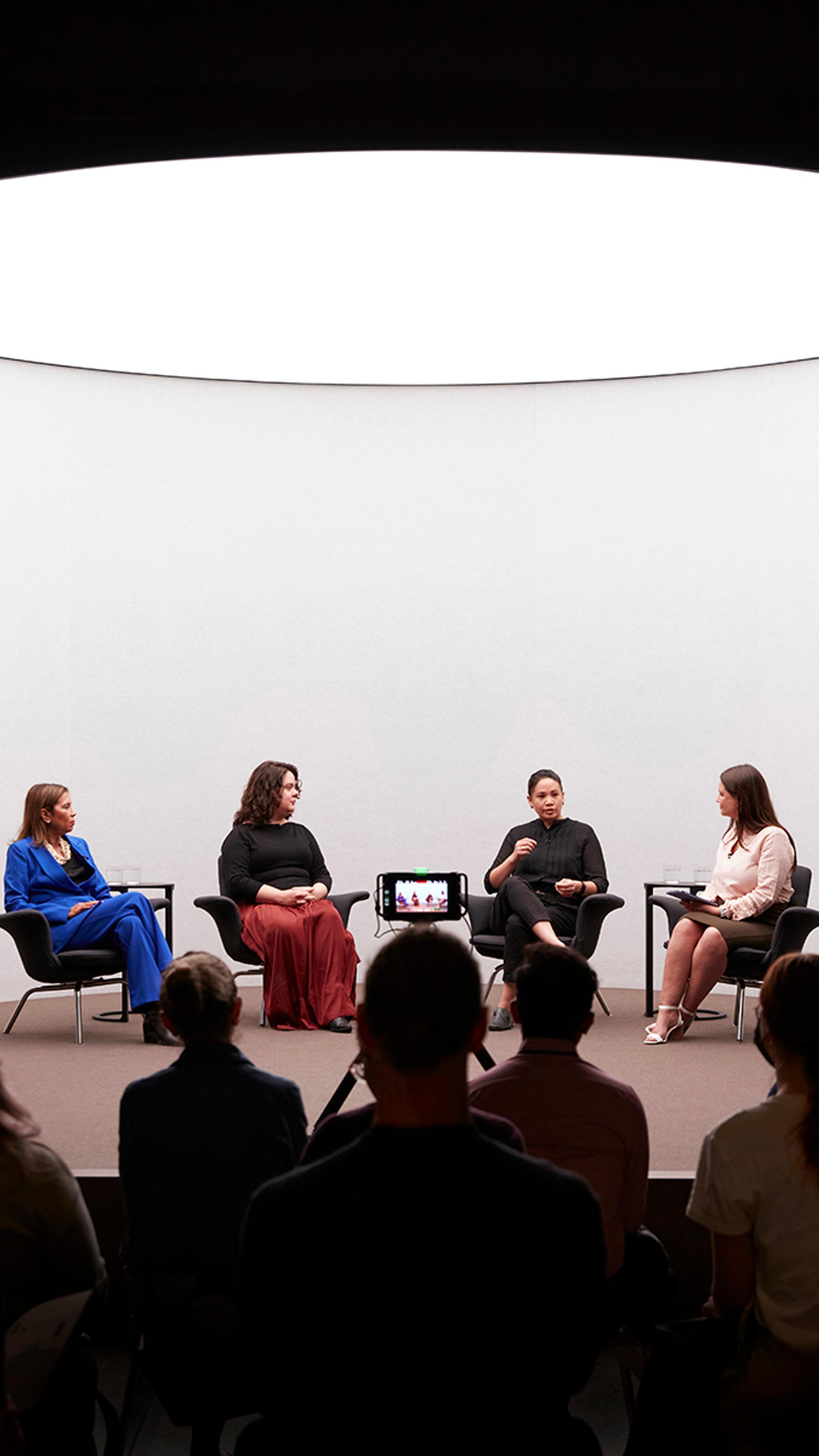 Four figures sit on a stage with a white background talking, there is a crowd watching the talk.