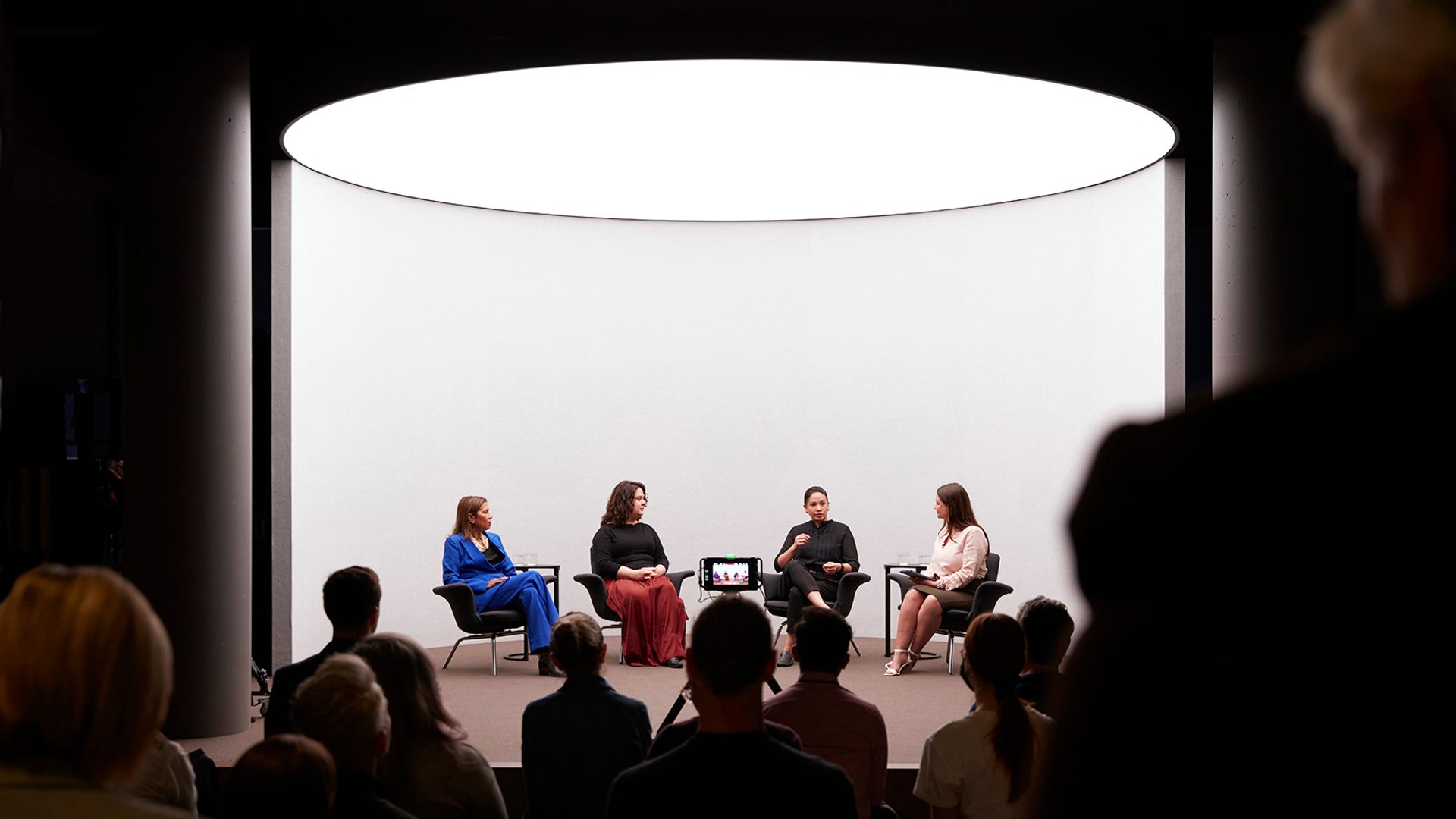 Four figures sit on a stage with a white background talking, there is a crowd watching the talk.