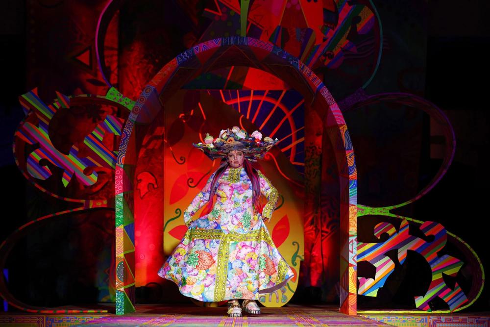 A model poses on the runway under a large archway, wearing a multicoloured floor-length dress and large hat.