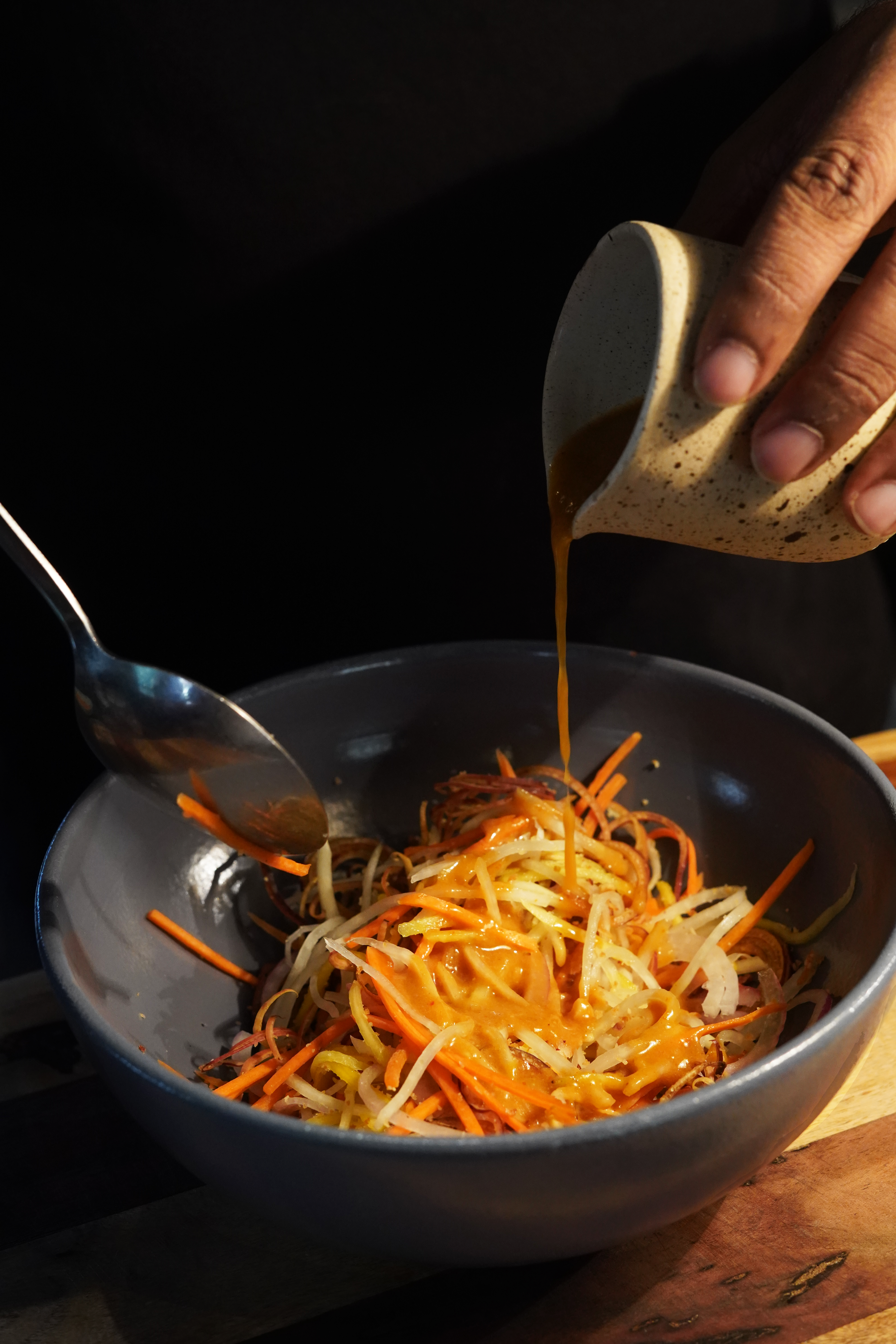 Dressing is poured from a small white vessel onto a salad in a blue bowl. The vessel is held by a brown hand.
