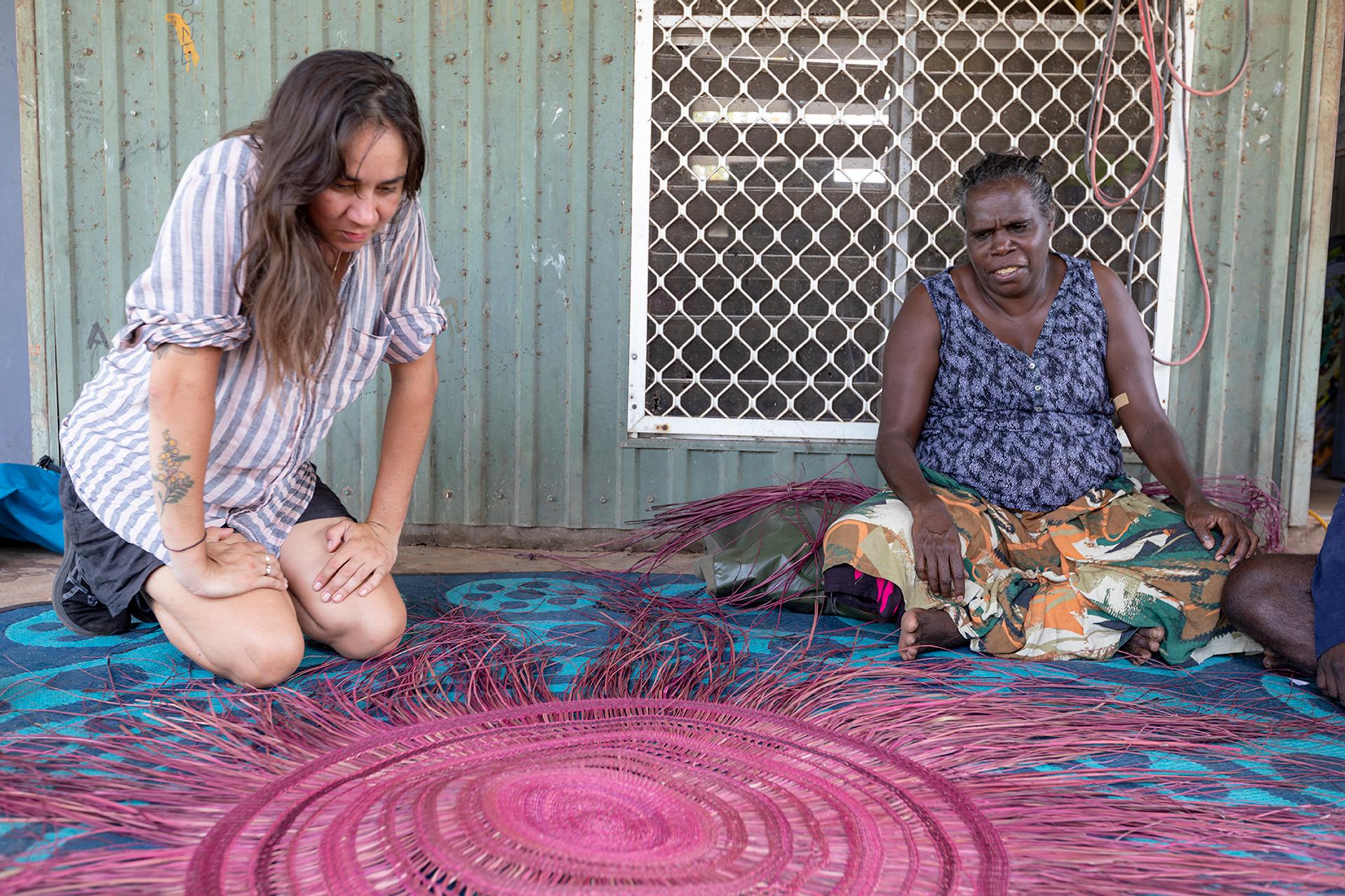 Two women sit outside on a blue patterned mat. The wall behind them is faded green and slightly rusted corrugated metal.
