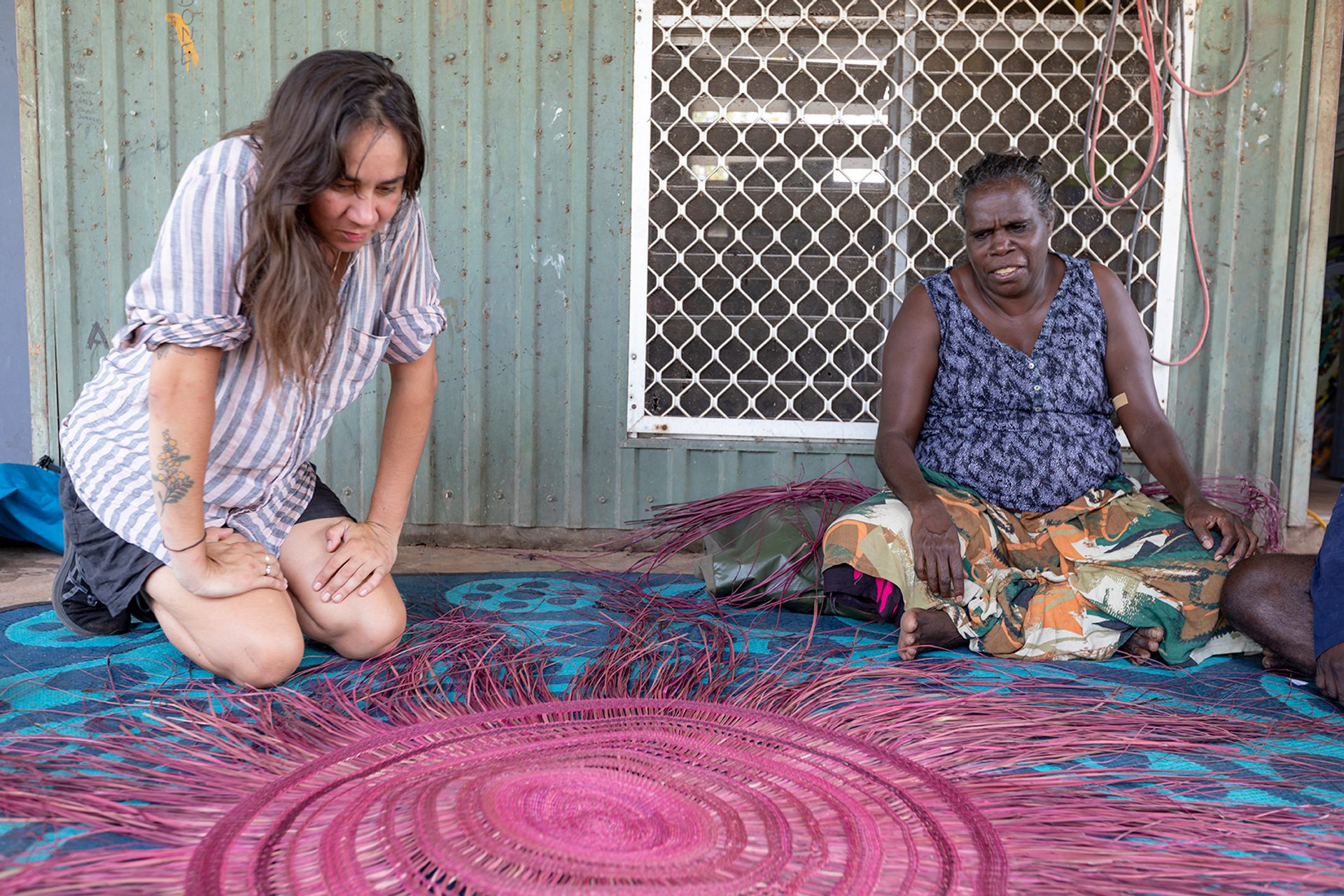 Two women sit outside on a blue patterned mat. The wall behind them is faded green and slightly rusted corrugated metal.