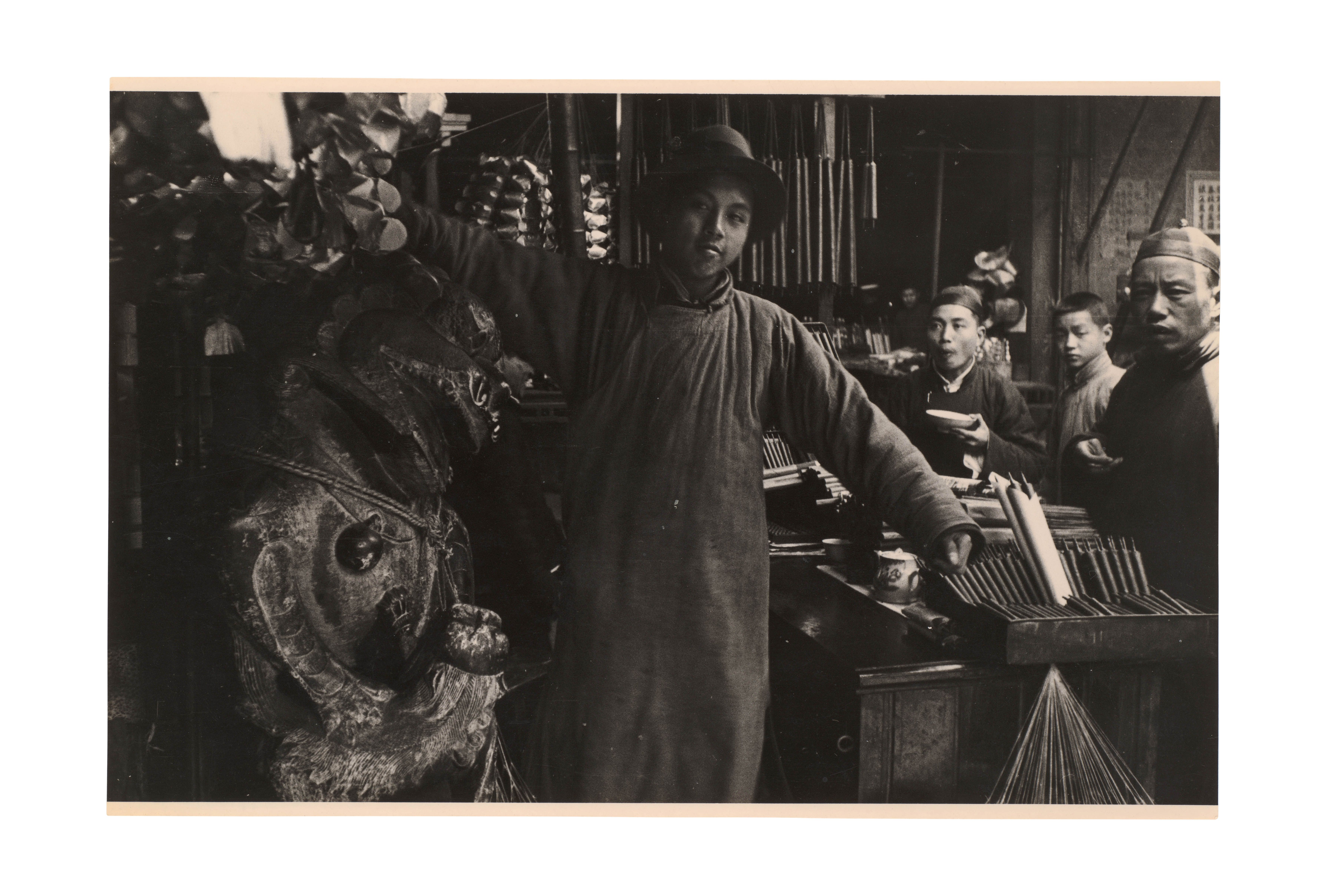 China around 1910–1914, a man holds up a traditional clothing piece in a market setting.