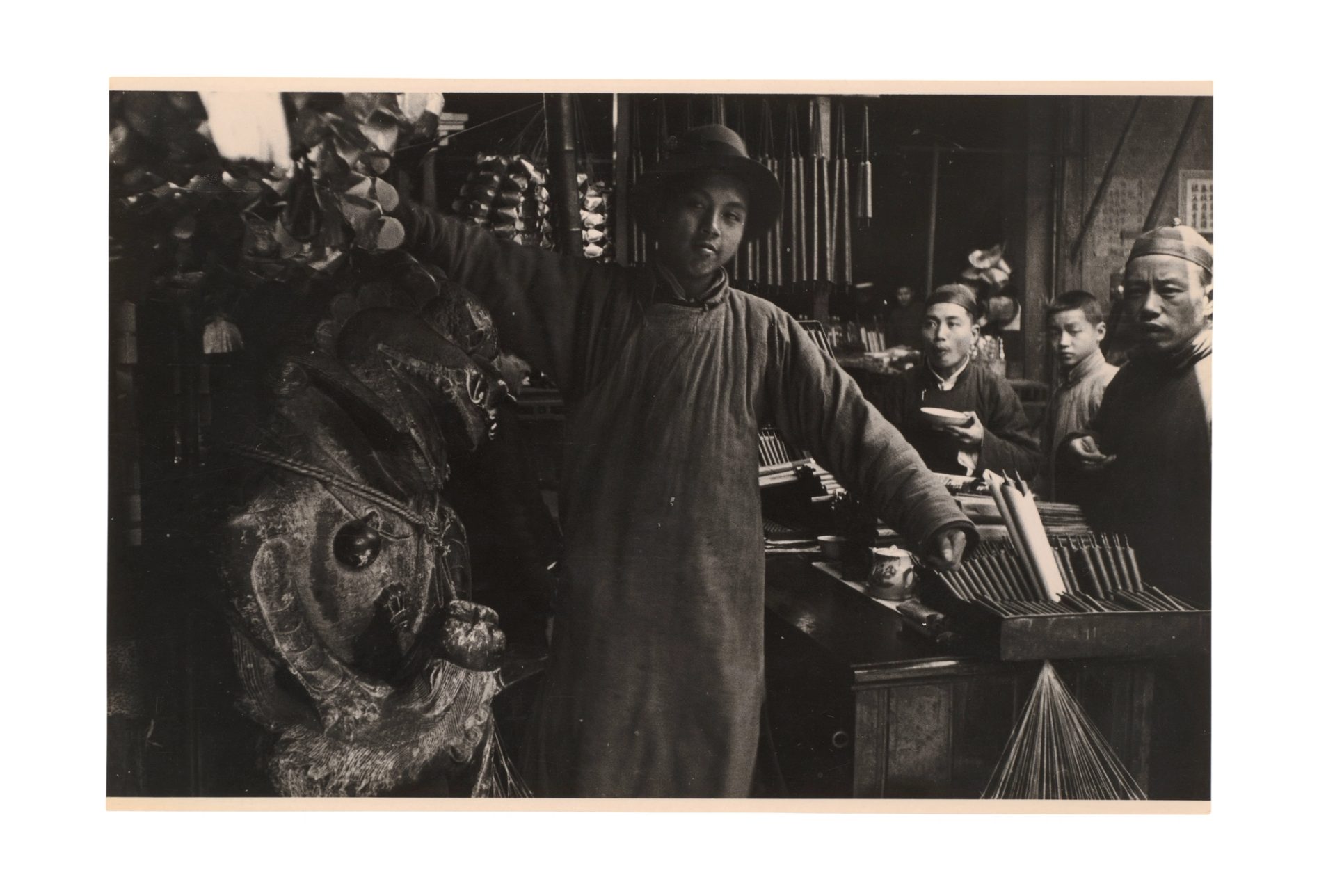 China around 1910–1914, a man holds up a traditional clothing piece in a market setting.