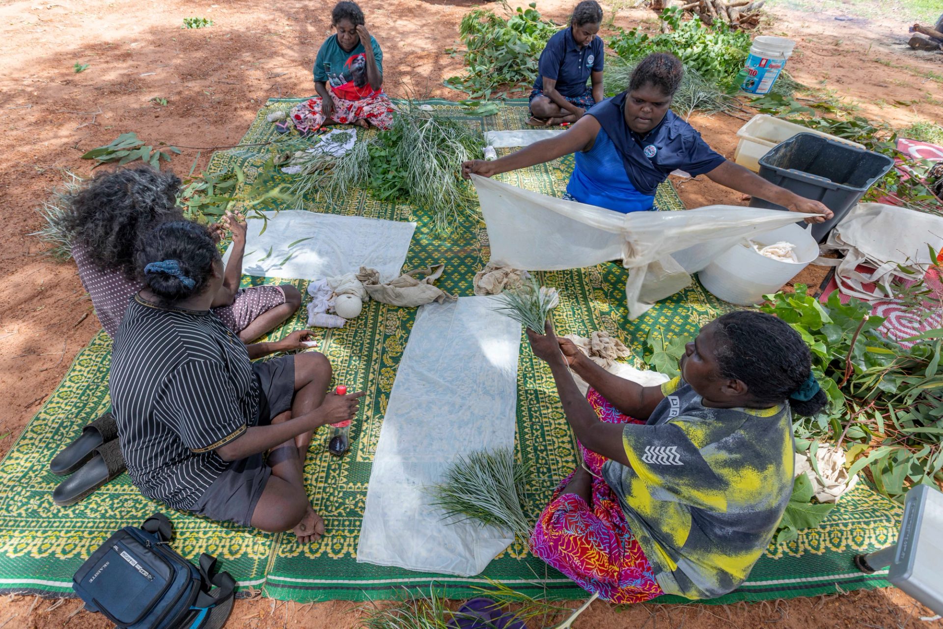 Six women sit outside on a large green and yellow mat. They are holding various plant clippings and pieces of white textiles.