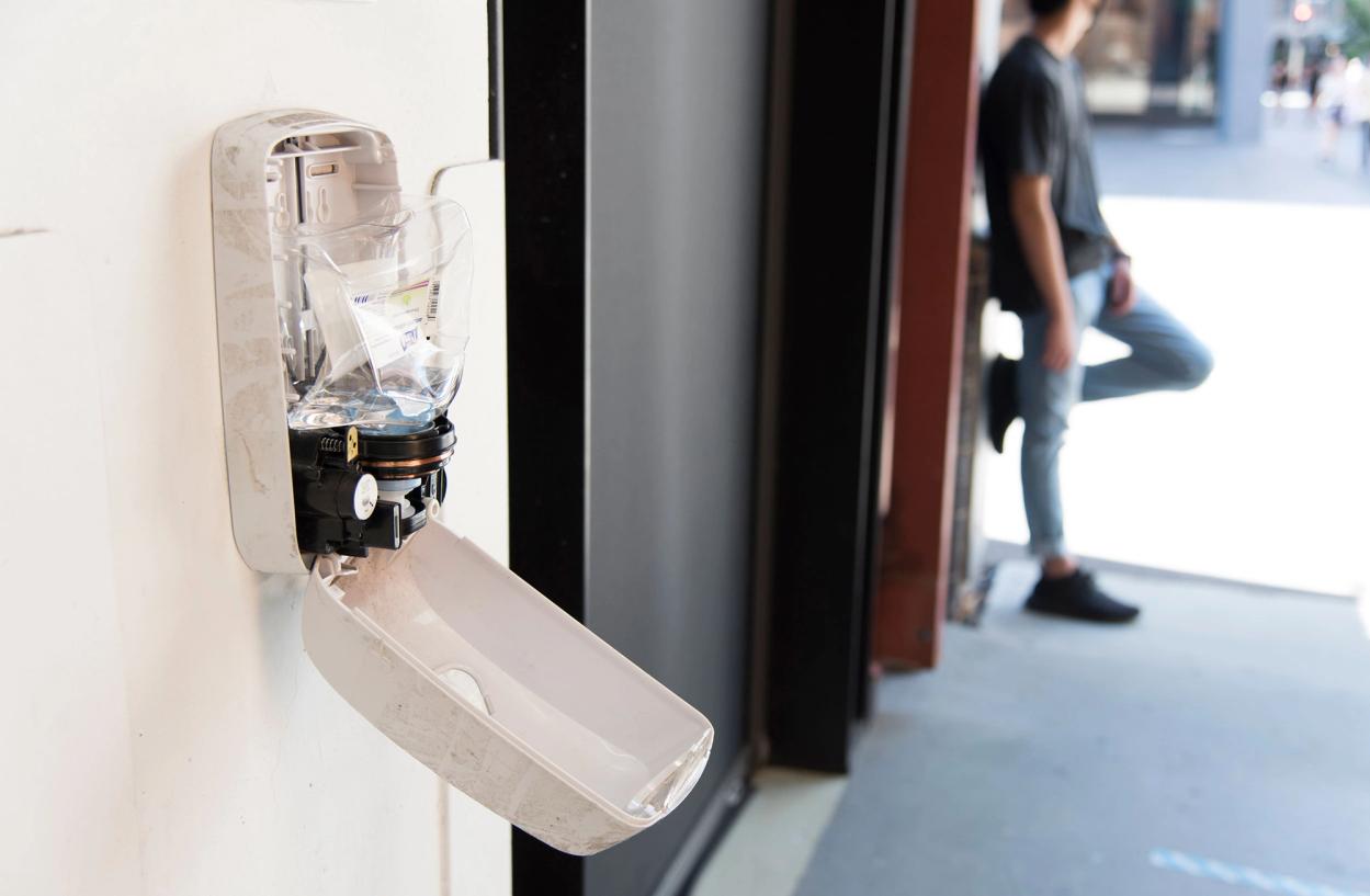 An open hand sanitiser dispenser on a white wall. A person leans against the same wall in the distance.