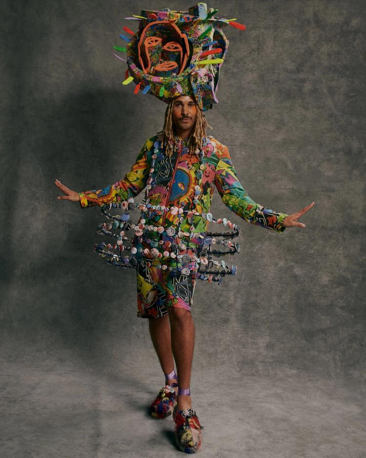 A model poses against a grey background wearing a multicoloured outfit and a circular wire frame structure adorned with buttons.