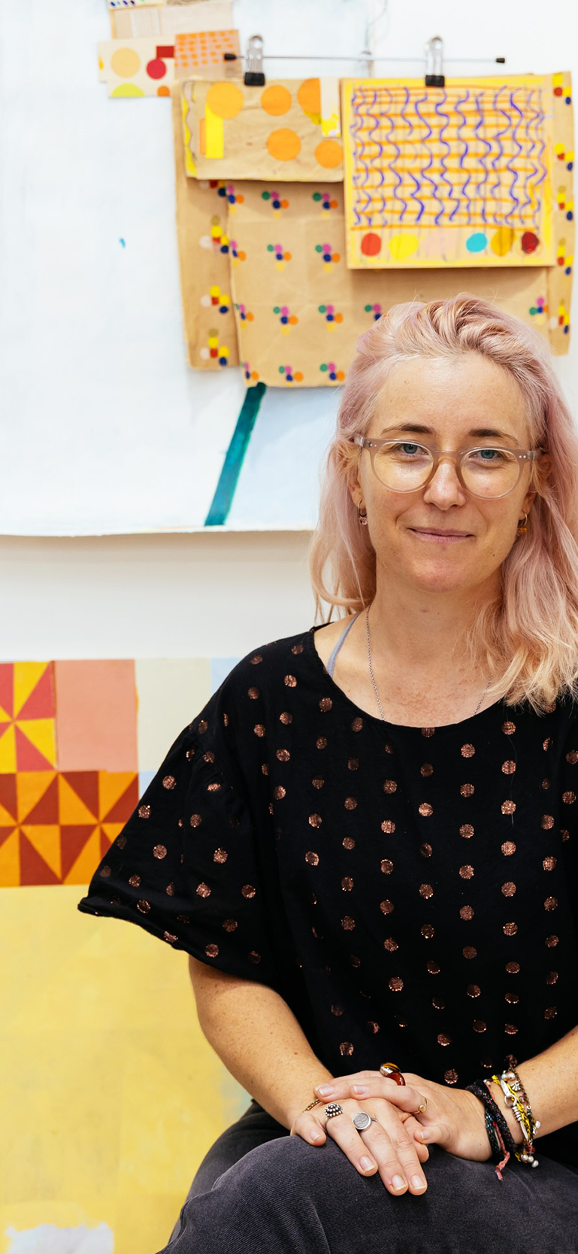 A woman with light pink hair wearing a spotted shirt and glasses sits surrounded by art materials in front of a wall of small and medium paper artwork.