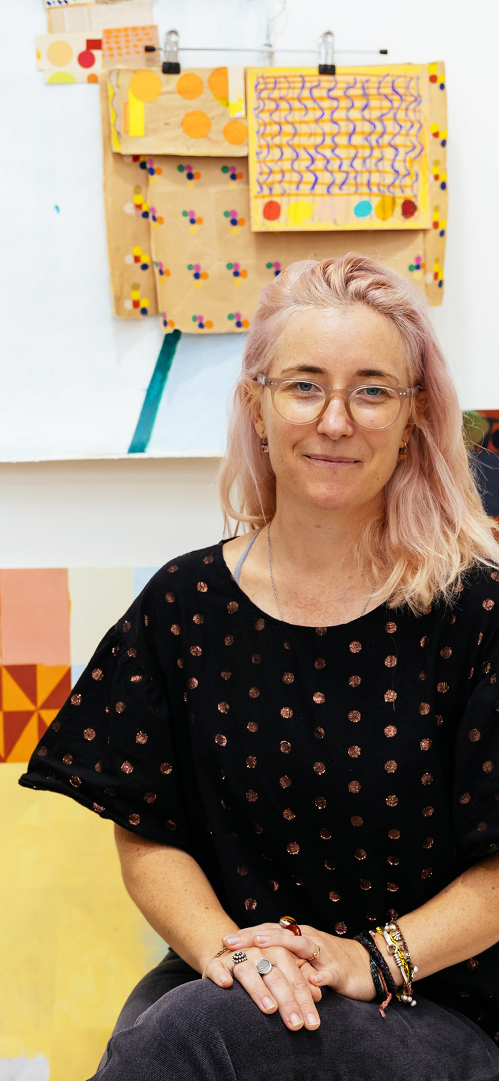 A woman with light pink hair wearing a spotted shirt and glasses sits surrounded by art materials in front of a wall of small and medium paper artwork.