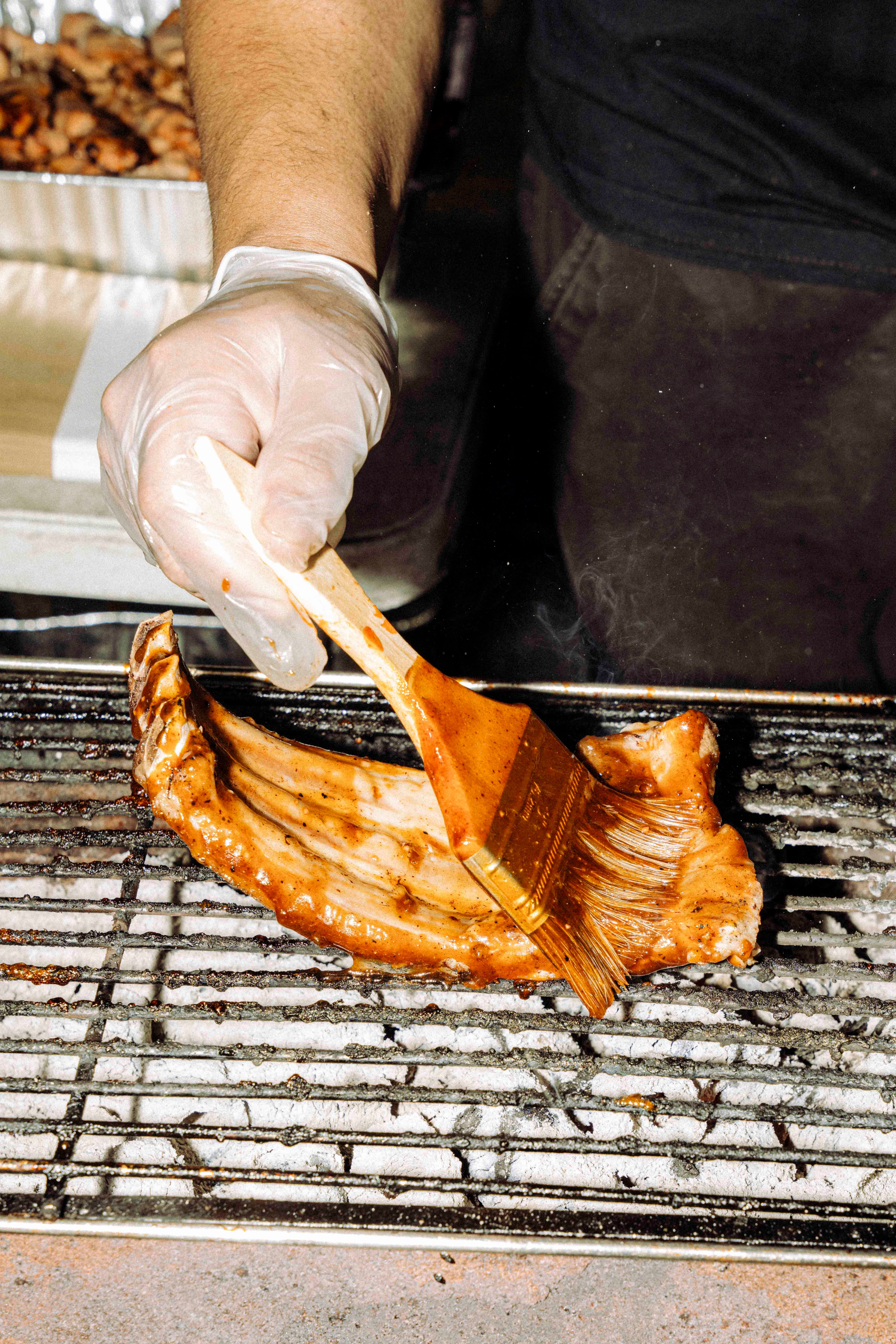 Close-up view of a black-attired street vendor in white plastic gloves who brushes glaze onto a large piece of bony meat that sits on the grill of a barbecue.