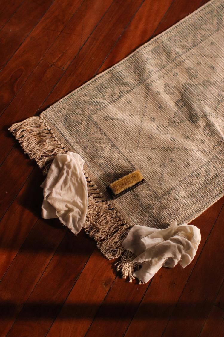 A small rug with tassels on polished wooden floorboards. On the rug is a shoe scrubbing brush and some cloth.