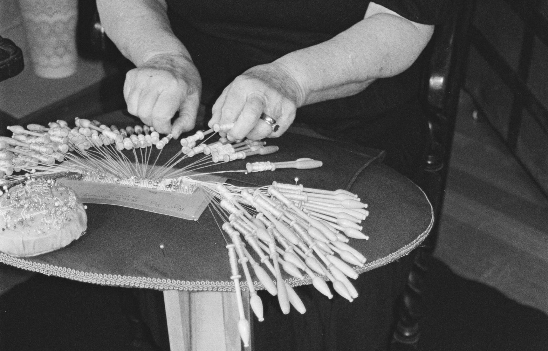 Black and white image of a person's hands (Barbara Stokes) holding the parts of a bobbin lacemaking machine. The machine is on a lace pillow table with pins pinned onto its surface.