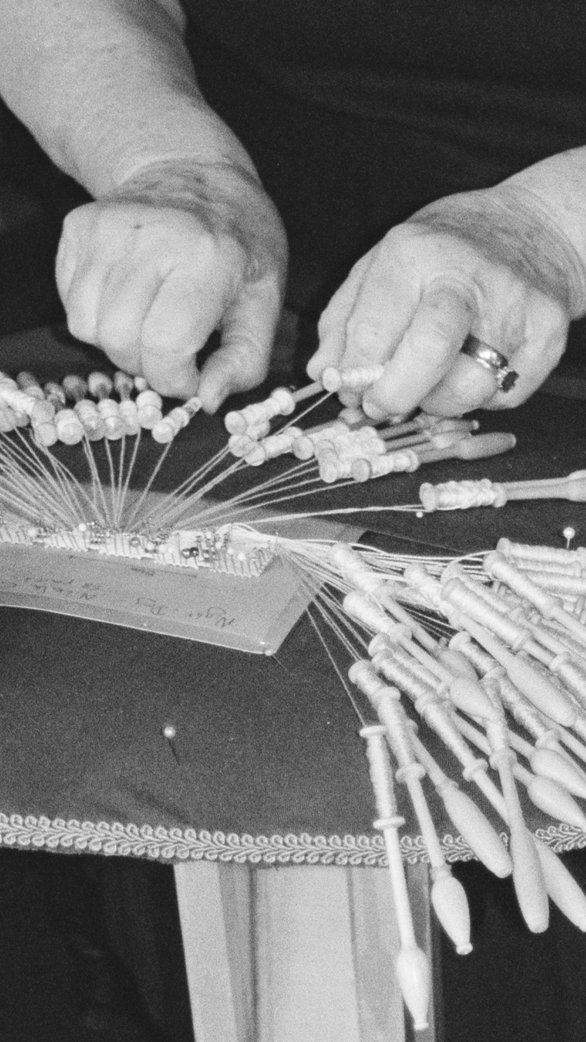 Black and white image of a person's hands (Barbara Stokes) holding the parts of a bobbin lacemaking machine. The machine is on a lace pillow table with pins pinned onto its surface.