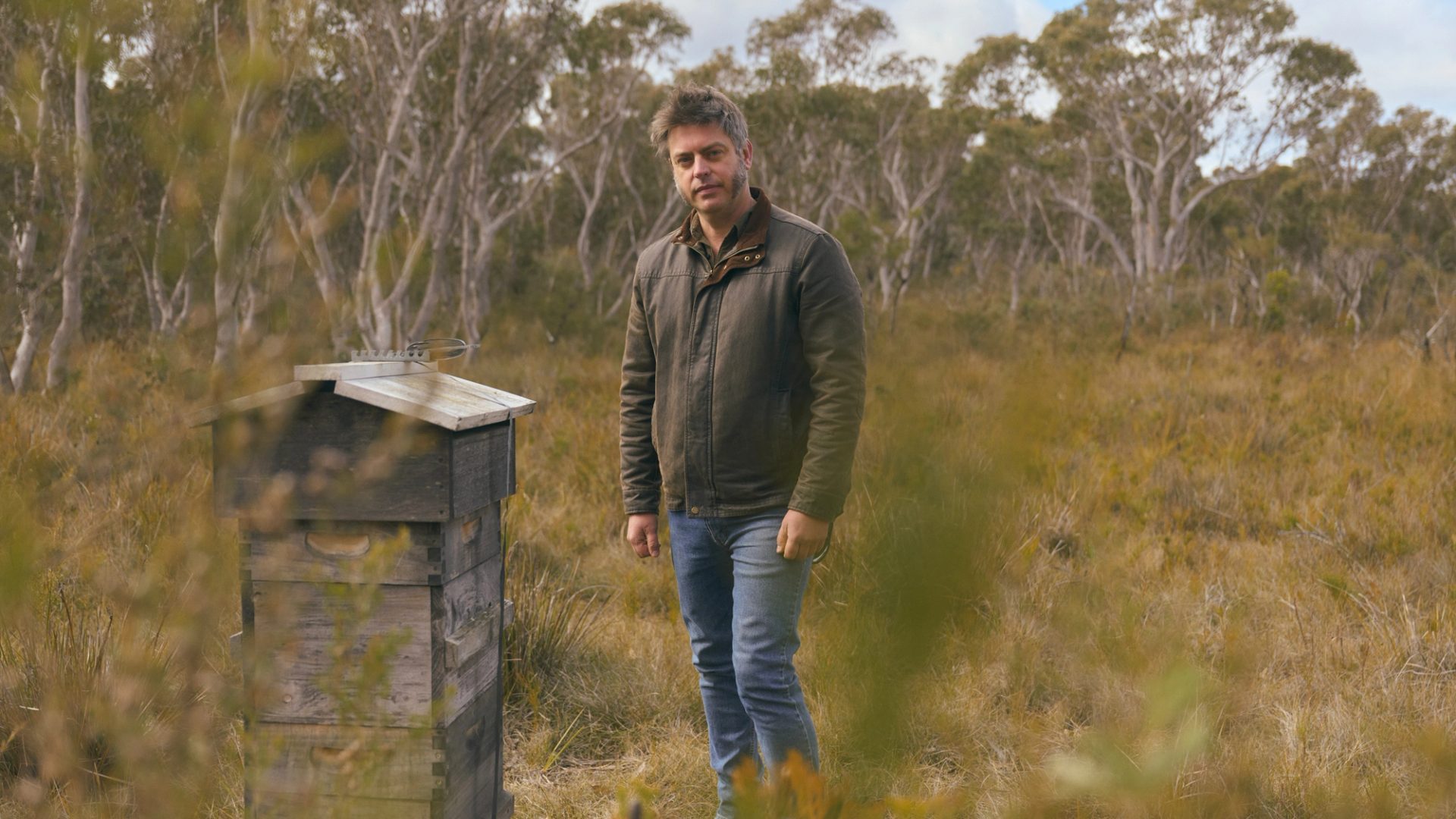 A man stands in a grassy, wooded area, next to a small, weathered wooden structure. He is wearing a brown jacket and jeans, looking directly at the camera with a calm expression. The background features tall trees with a cloudy sky above, creating a serene, natural atmosphere.
