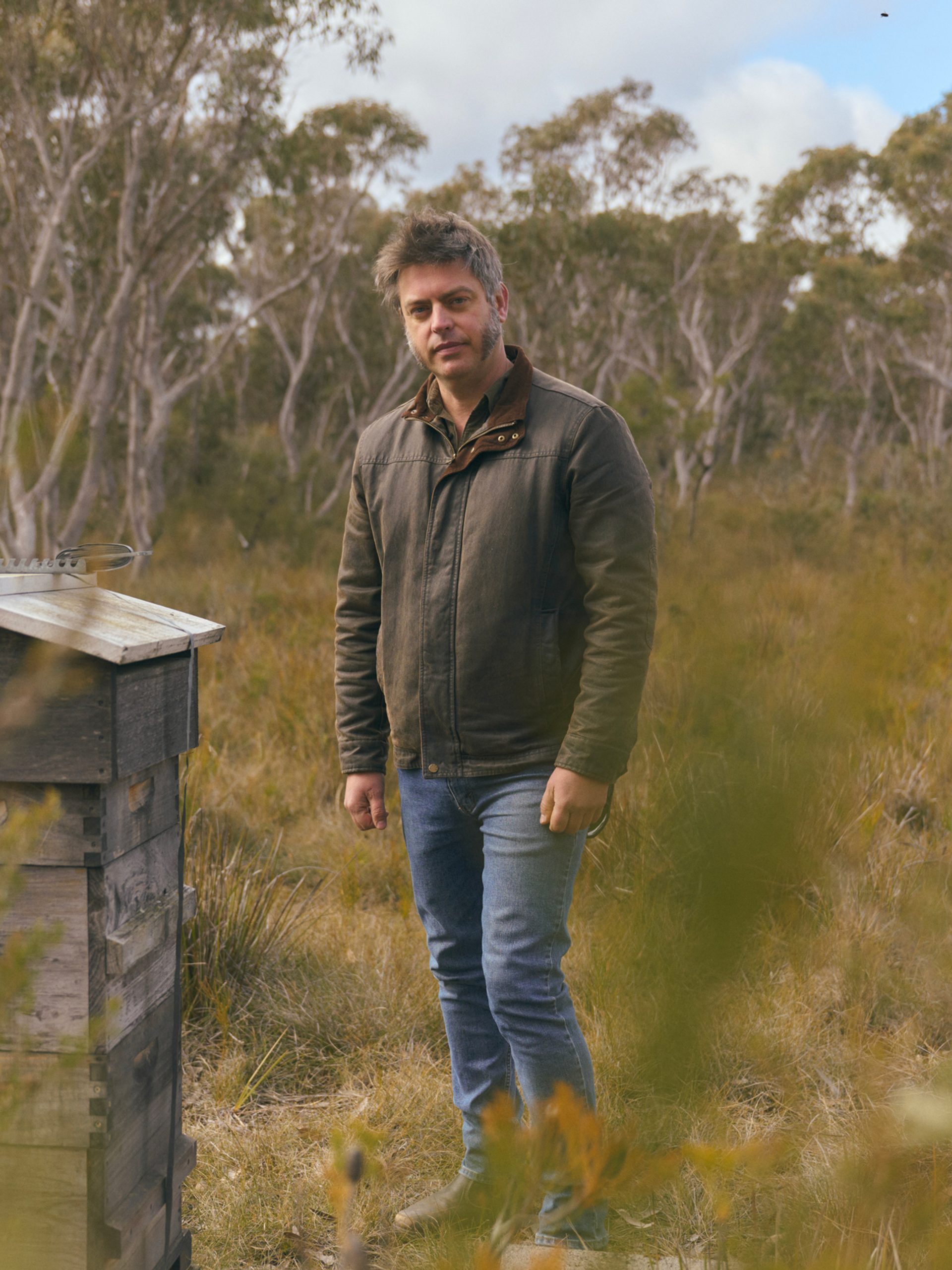 A man stands in a grassy, wooded area, next to a small, weathered wooden structure. He is wearing a brown jacket and jeans, looking directly at the camera with a calm expression. The background features tall trees with a cloudy sky above, creating a serene, natural atmosphere.