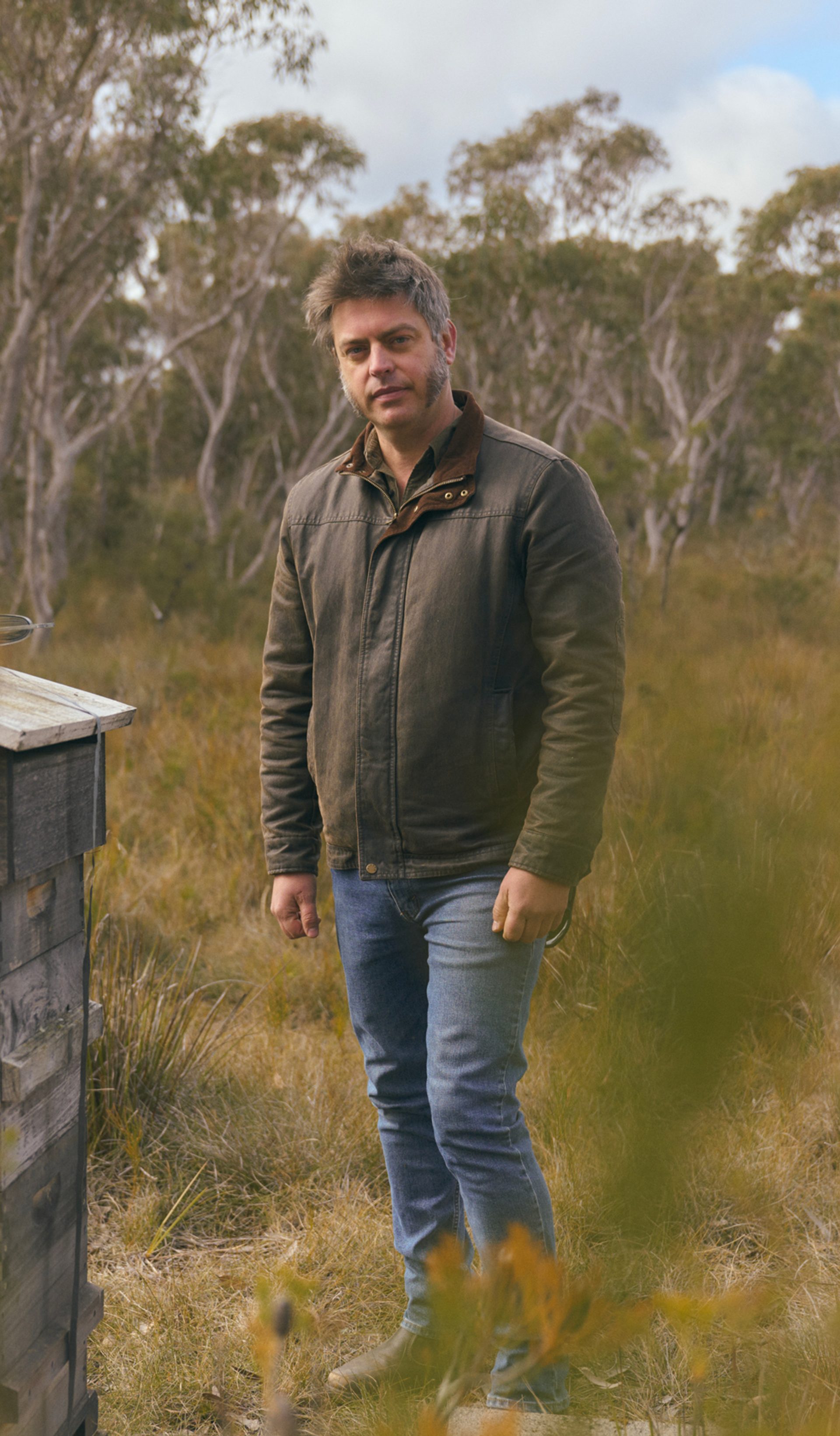 A man stands in a grassy, wooded area, next to a small, weathered wooden structure. He is wearing a brown jacket and jeans, looking directly at the camera with a calm expression. The background features tall trees with a cloudy sky above, creating a serene, natural atmosphere.