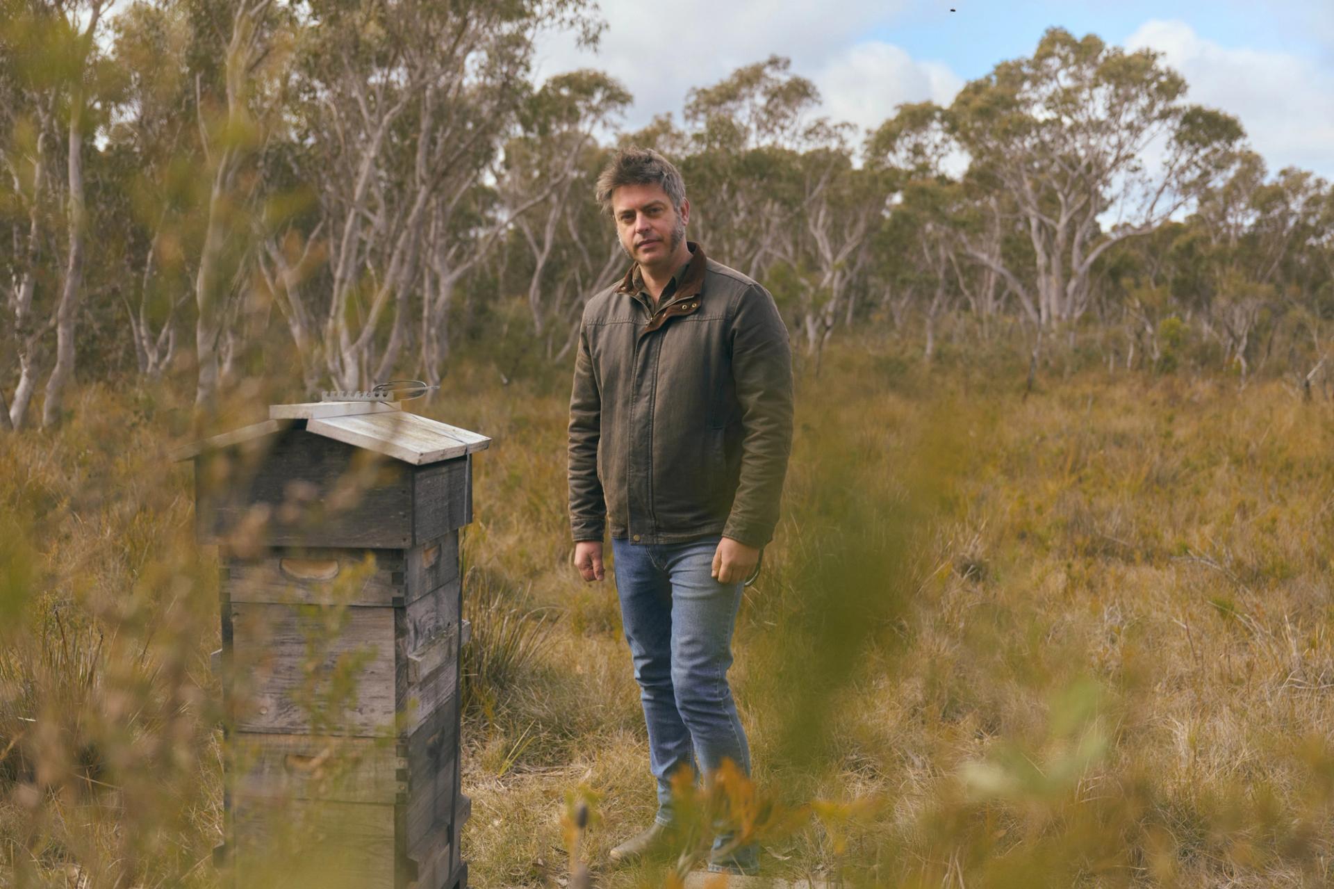 A man stands in a grassy, wooded area, next to a small, weathered wooden structure. He is wearing a brown jacket and jeans, looking directly at the camera with a calm expression. The background features tall trees with a cloudy sky above, creating a serene, natural atmosphere.