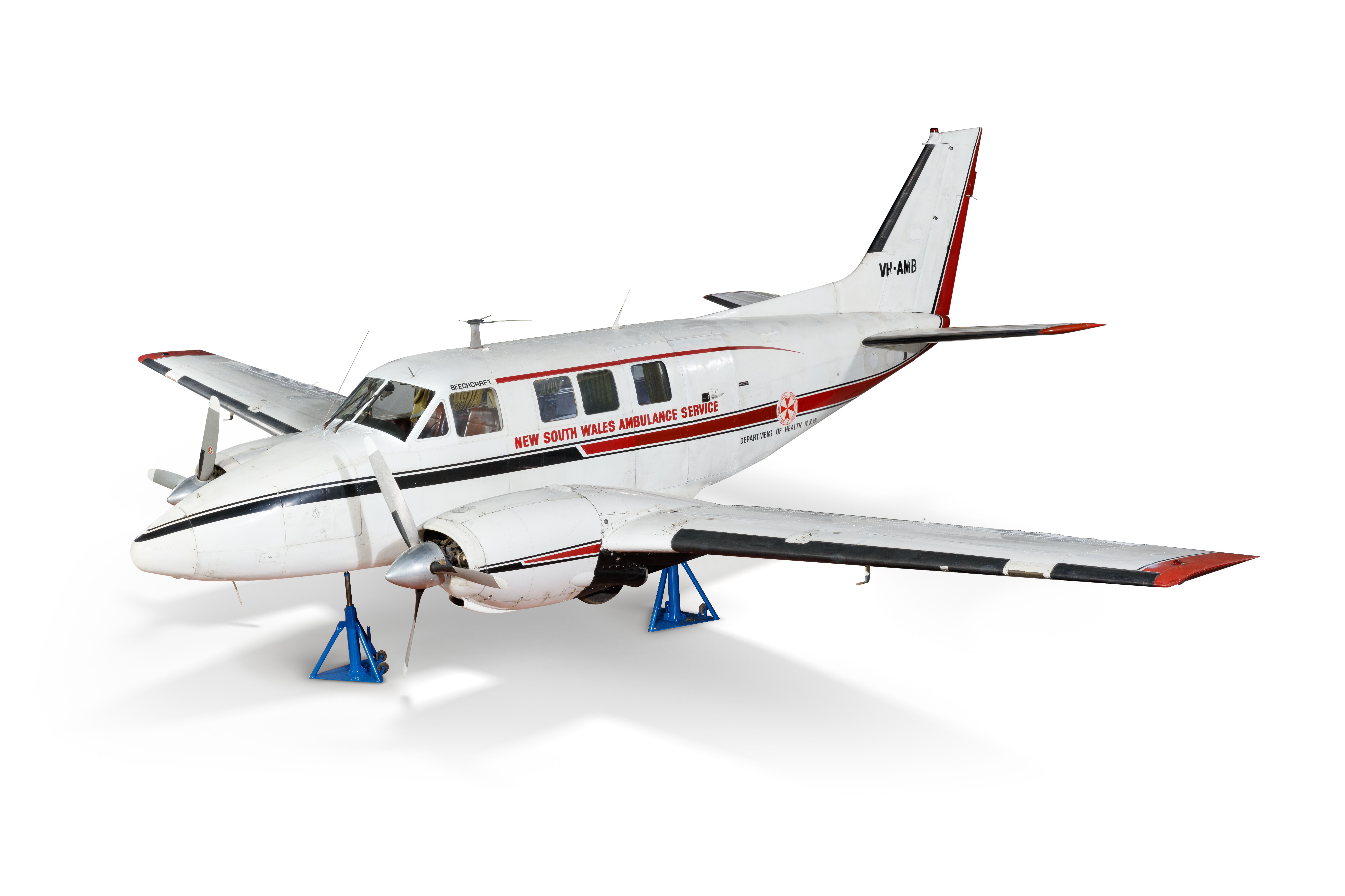 Angled side view of a white twin propeller airplane sitting on blue triangular supports. 'New South Wales Ambulance Service' is written on the side.