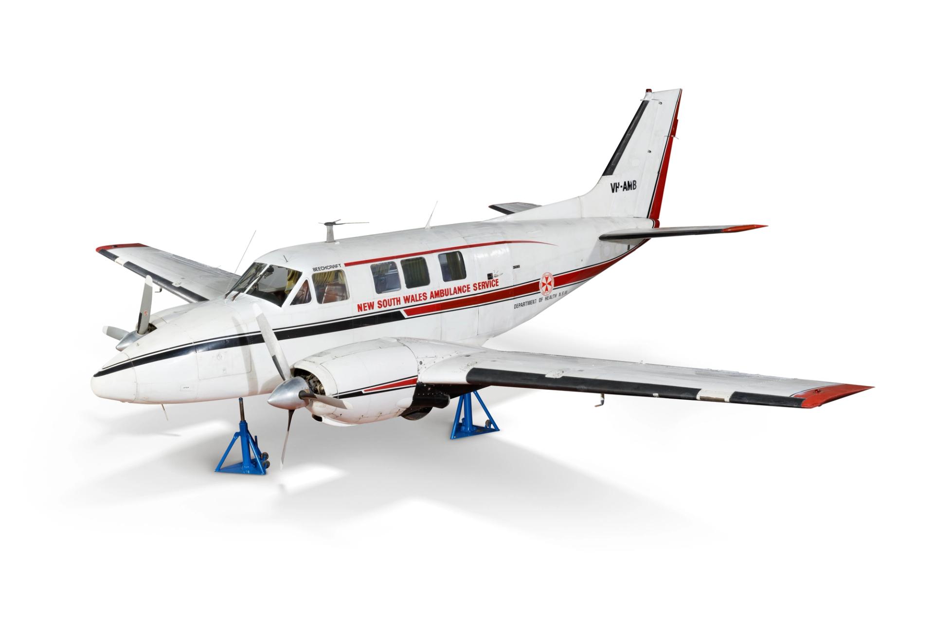 Angled side view of a white twin propeller airplane sitting on blue triangular supports. 'New South Wales Ambulance Service' is written on the side.
