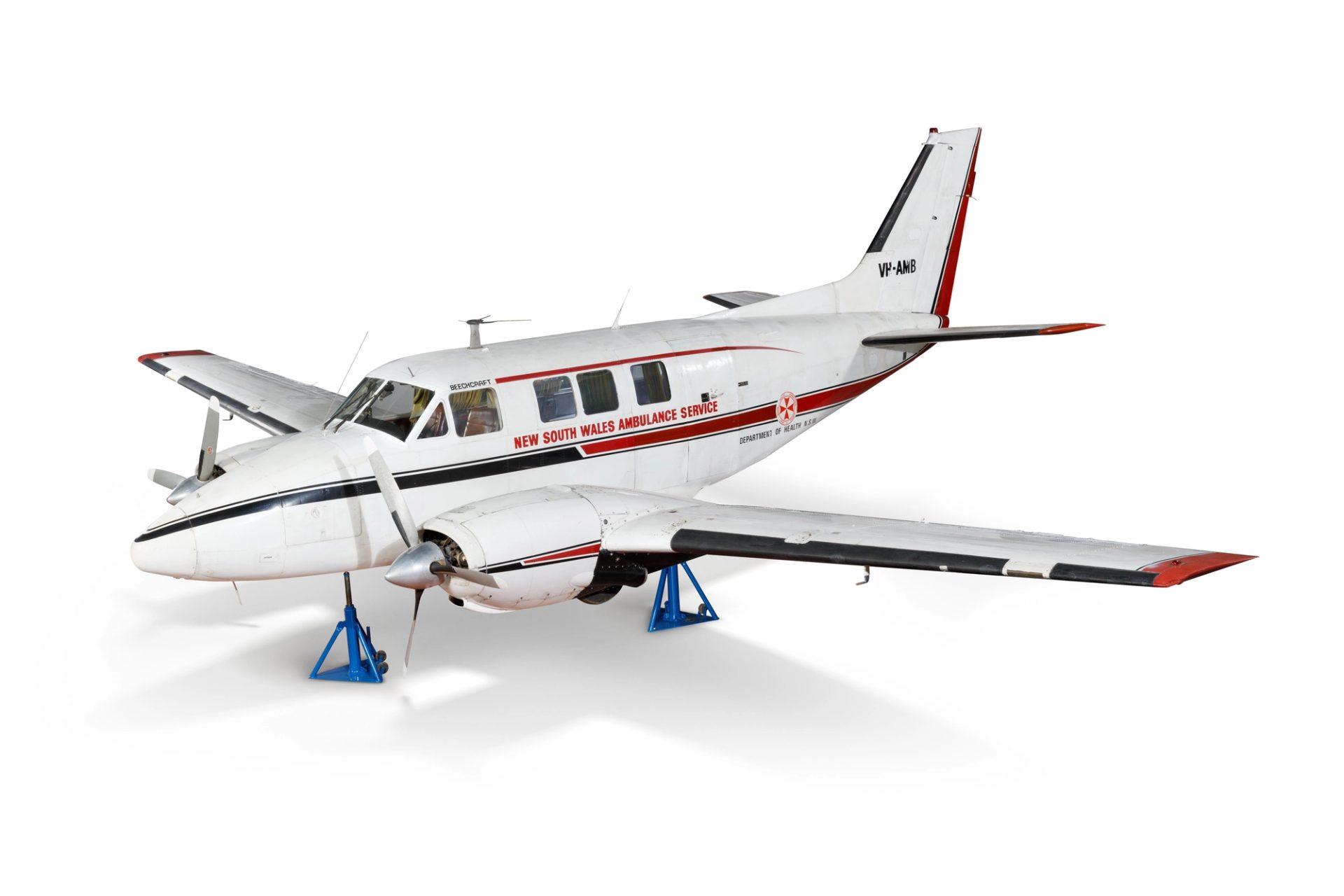 Angled side view of a white twin propeller airplane sitting on blue triangular supports. 'New South Wales Ambulance Service' is written on the side.