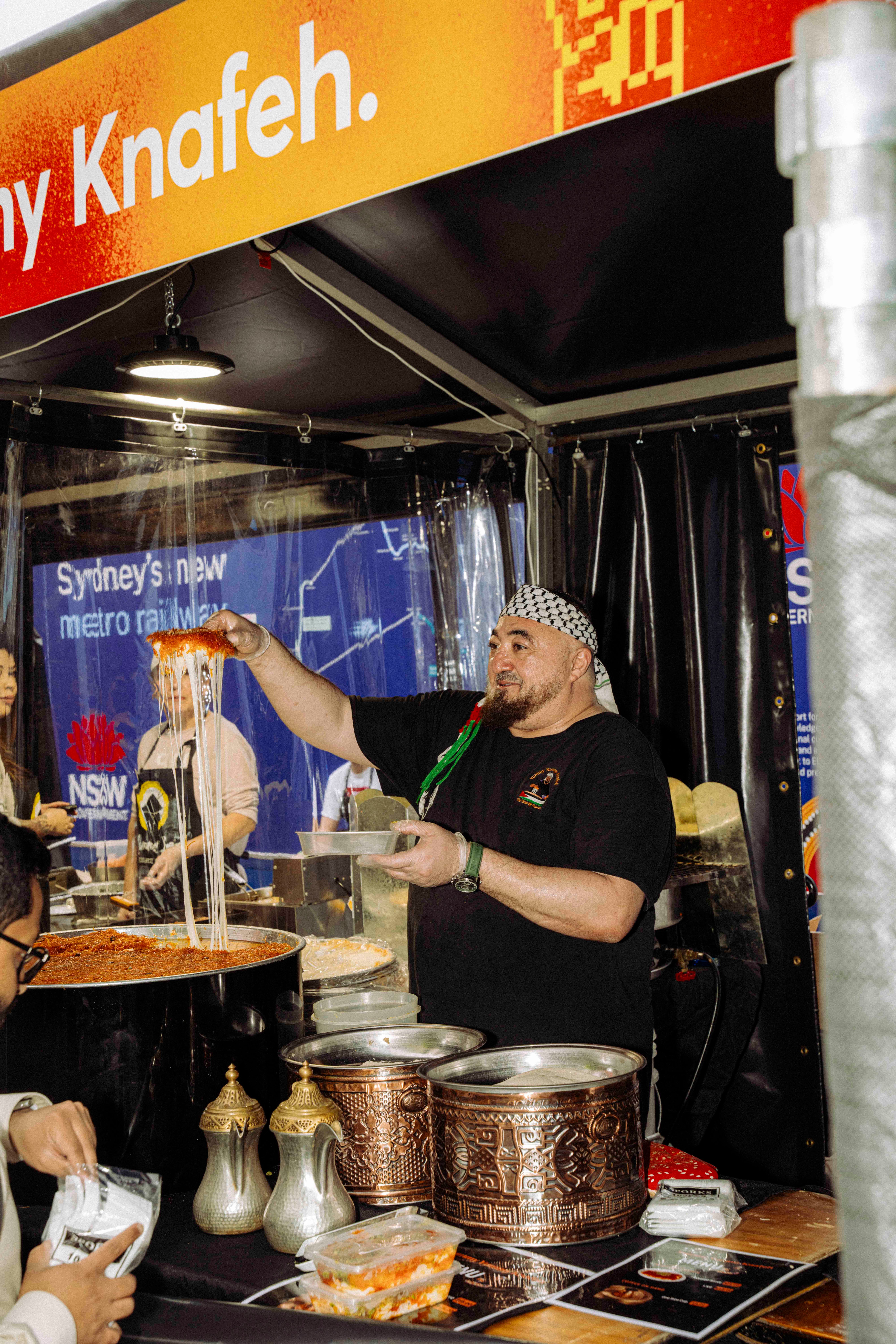 A smiling vendor in a patterned headscarf pulls golden strands from a large pot at a food stall, with colourful signage in the background that reads ‘Knafeh’.
