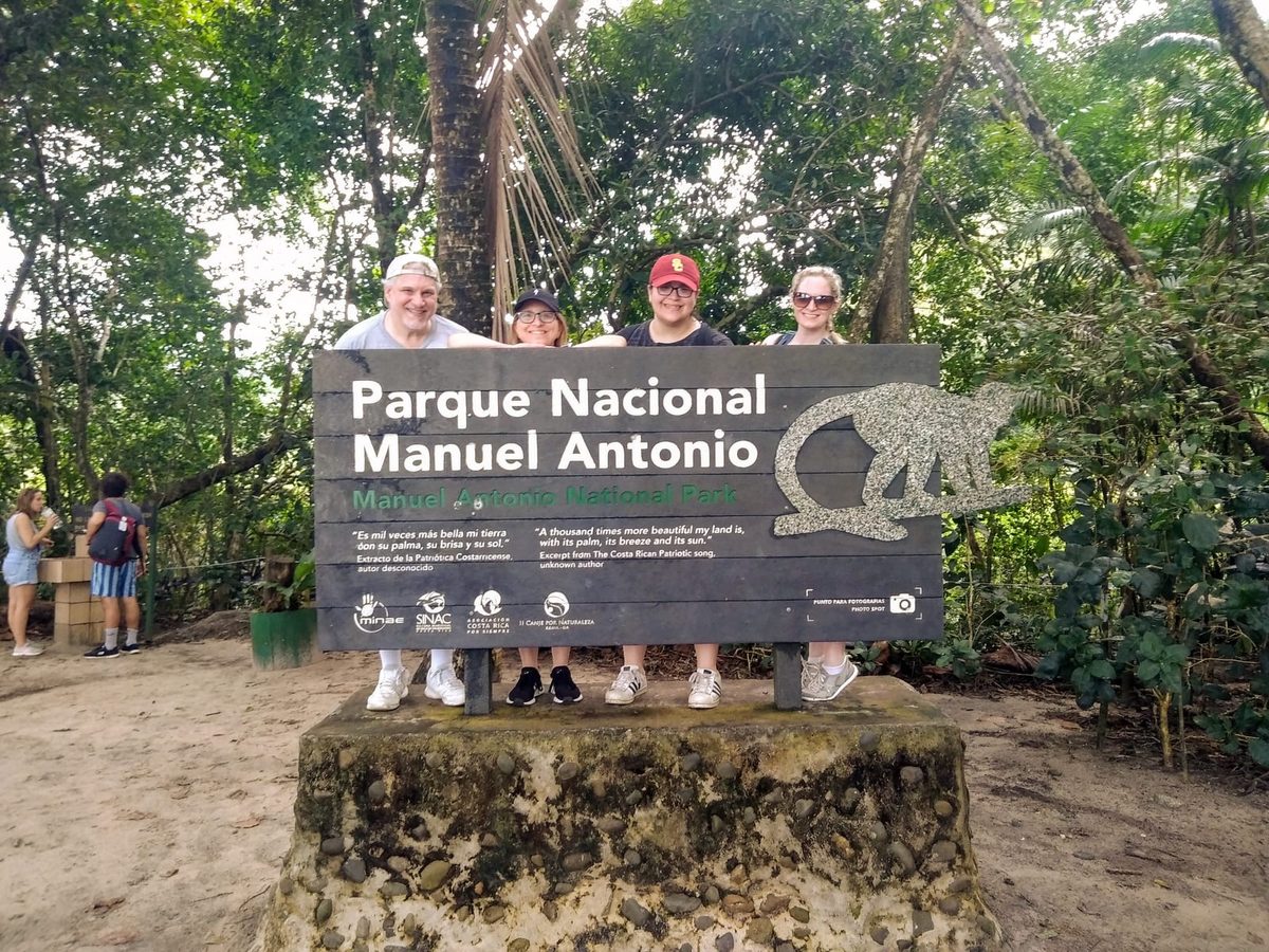 Four visitors standing behind the dark wooden Parque Nacional Manuel Antonio sign at the park entrance, jungle rising behind them