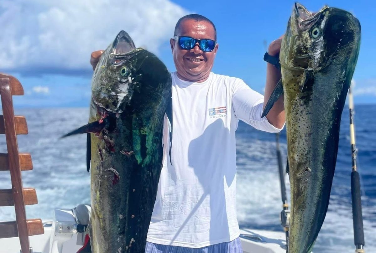 A man in sunglasses on the back deck of a fishing boat holding up a mahi-mahi in each hand, open sea behind him