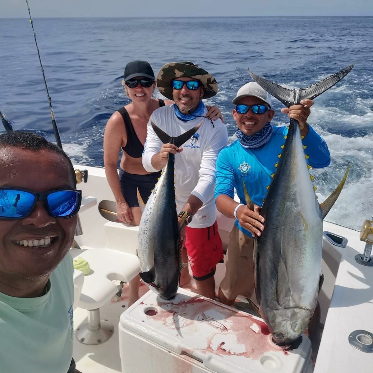 Two yellowfin tuna held up by their tails over the fish box on the back deck of a sportfishing boat, four people smiling in a selfie around them