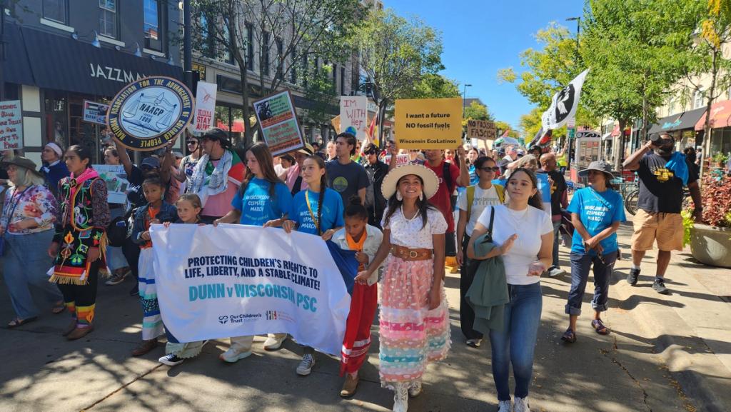 Marchers on a Wisconsin street carry a banner reading “Protecting children’s rights to life, liberty, and a stable climate — Dunn v. Wisconsin PSC”.