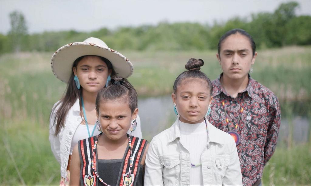 our young Indigenous youth standing together outdoors in a row, looking seriously at the camera with a grassy Wisconsin landscape and a body of water blurred in the background.