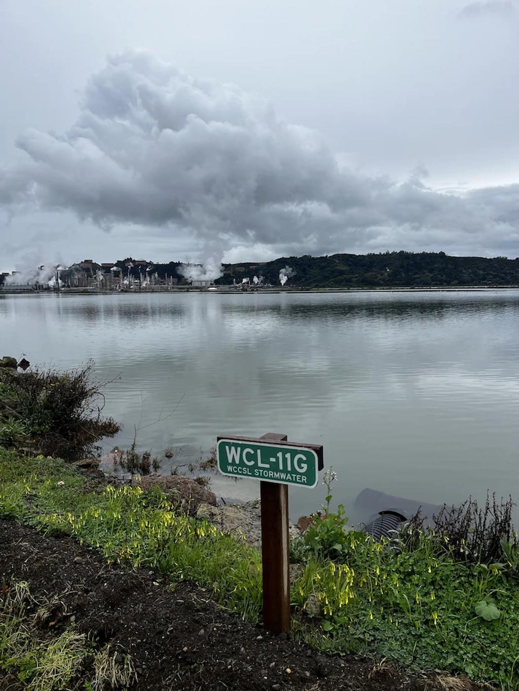 A monitoring-station sign on the bay shore under grey skies.