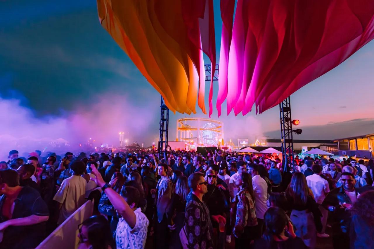 A crowd dancing at dusk under huge orange and pink fabric sails, a lit Ferris wheel behind them.