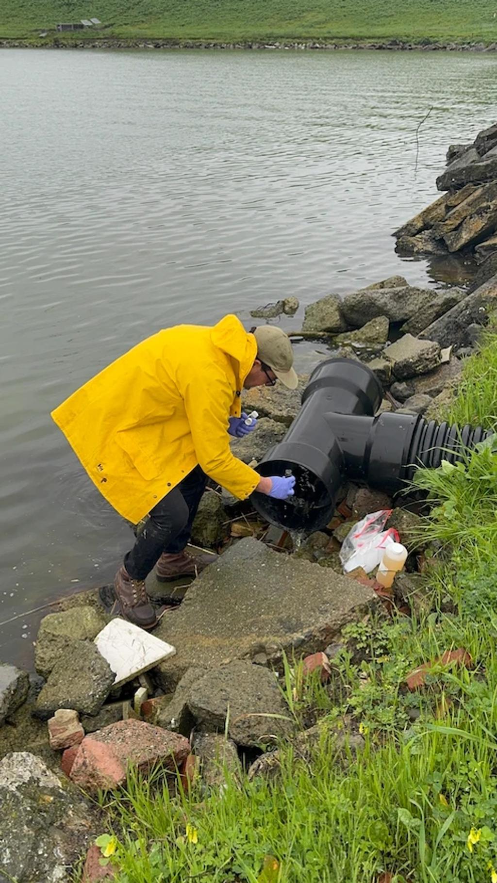 A field technician in a yellow raincoat sampling water from an outfall pipe.