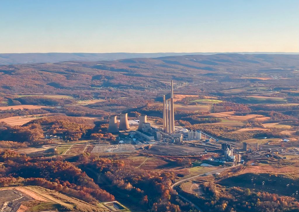 An aerial wide shot of a large industrial power plant with massive cooling towers and tall smokestacks, surrounded by rolling hills and rural landscape in autumn.