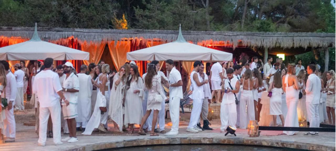 Guests dressed in white mingle by a pool under parasols and a lantern-lit thatched pavilion.