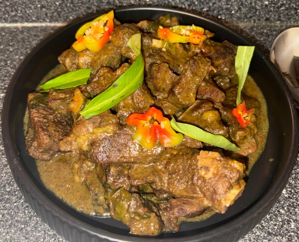 Authentic slow-braised Tarragon Curry Goat by Chef Kimberly Kee of Saucy Kim K, served family-style for luxury catering events. Features fresh tarragon infusion and Scotch Bonnet garnishes in a professional black ceramic bowl. Serving private events and group celebrations in Newport RI, Greenwich CT, and Boston MA.