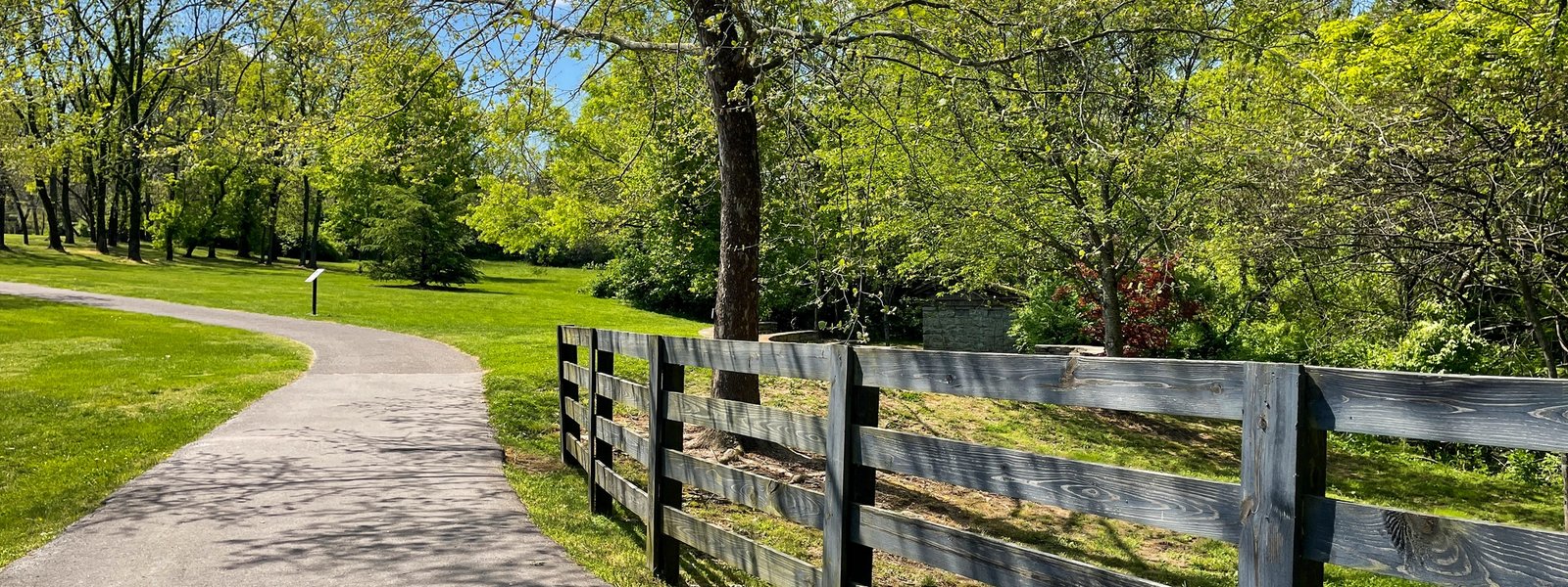 A quiet paved path curves alongside a wooden ranch fence and green trees on a spring day in Brentwood.