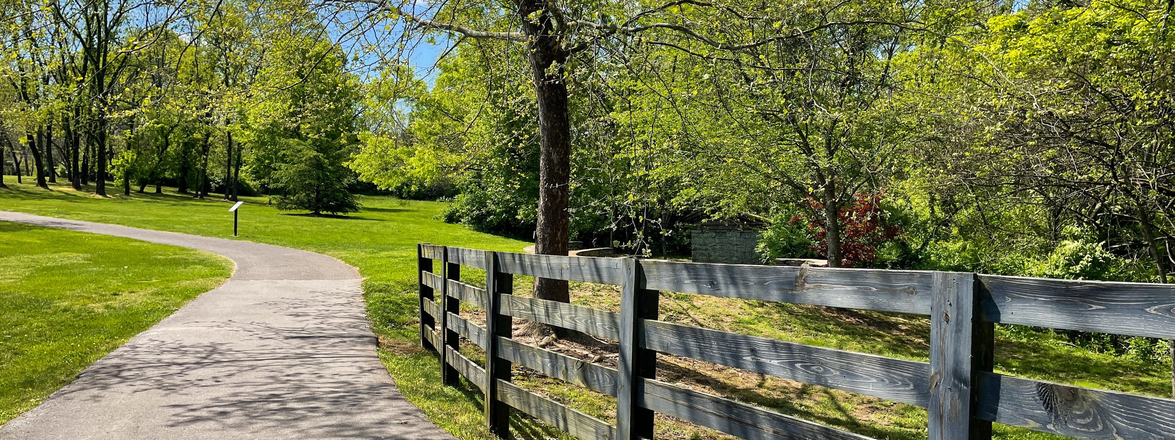 A quiet paved path curves alongside a wooden ranch fence and green trees on a spring day in Brentwood.