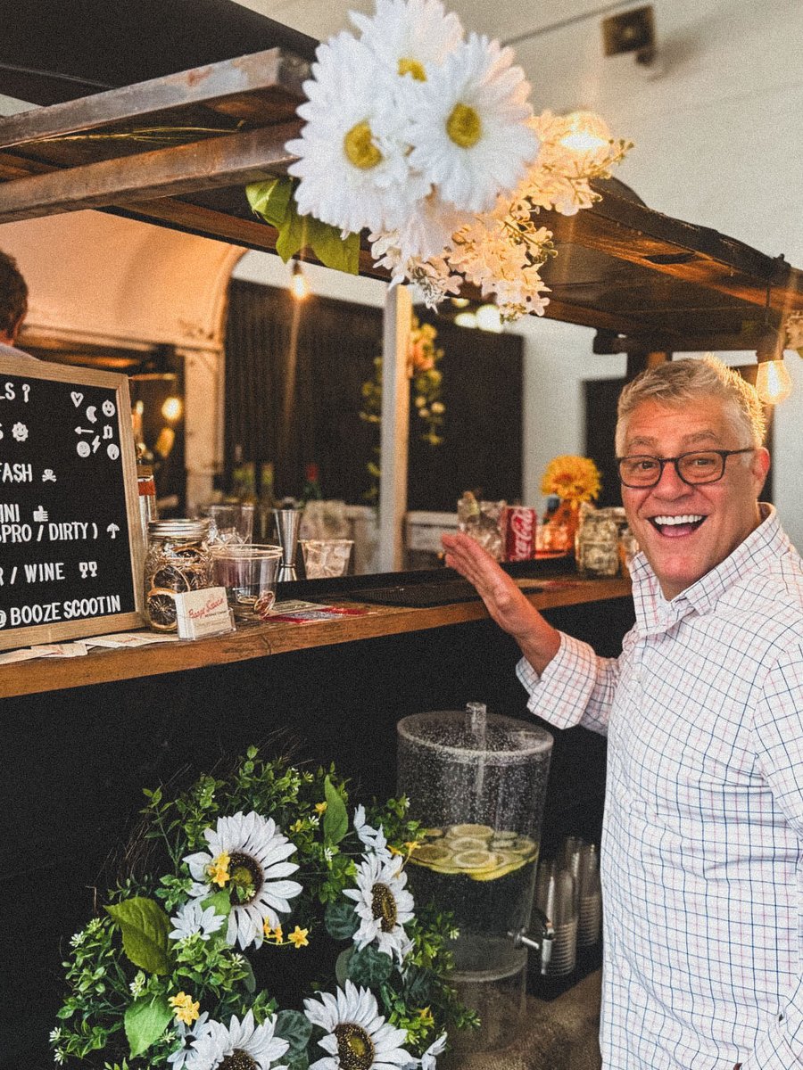A smiling man in a checked shirt points at the trailer's cocktail menu, sunflowers along the bar.