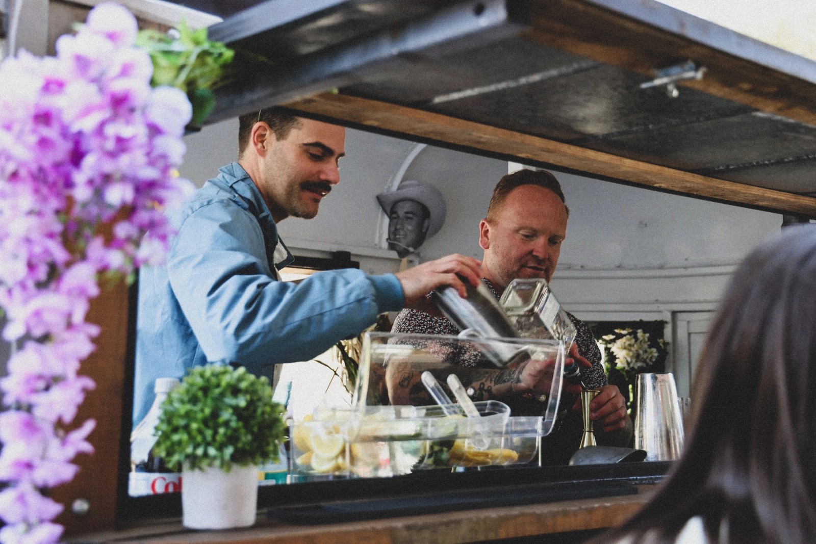 Two bartenders mixing drinks inside the Booze Scootin' trailer, lemons and mint on the bar in front of them.