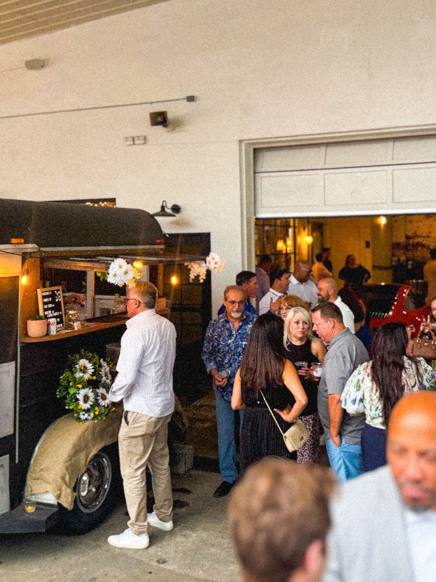 Guests hold cocktails around the Howdy trailer parked inside a venue, sunflowers by the service window.
