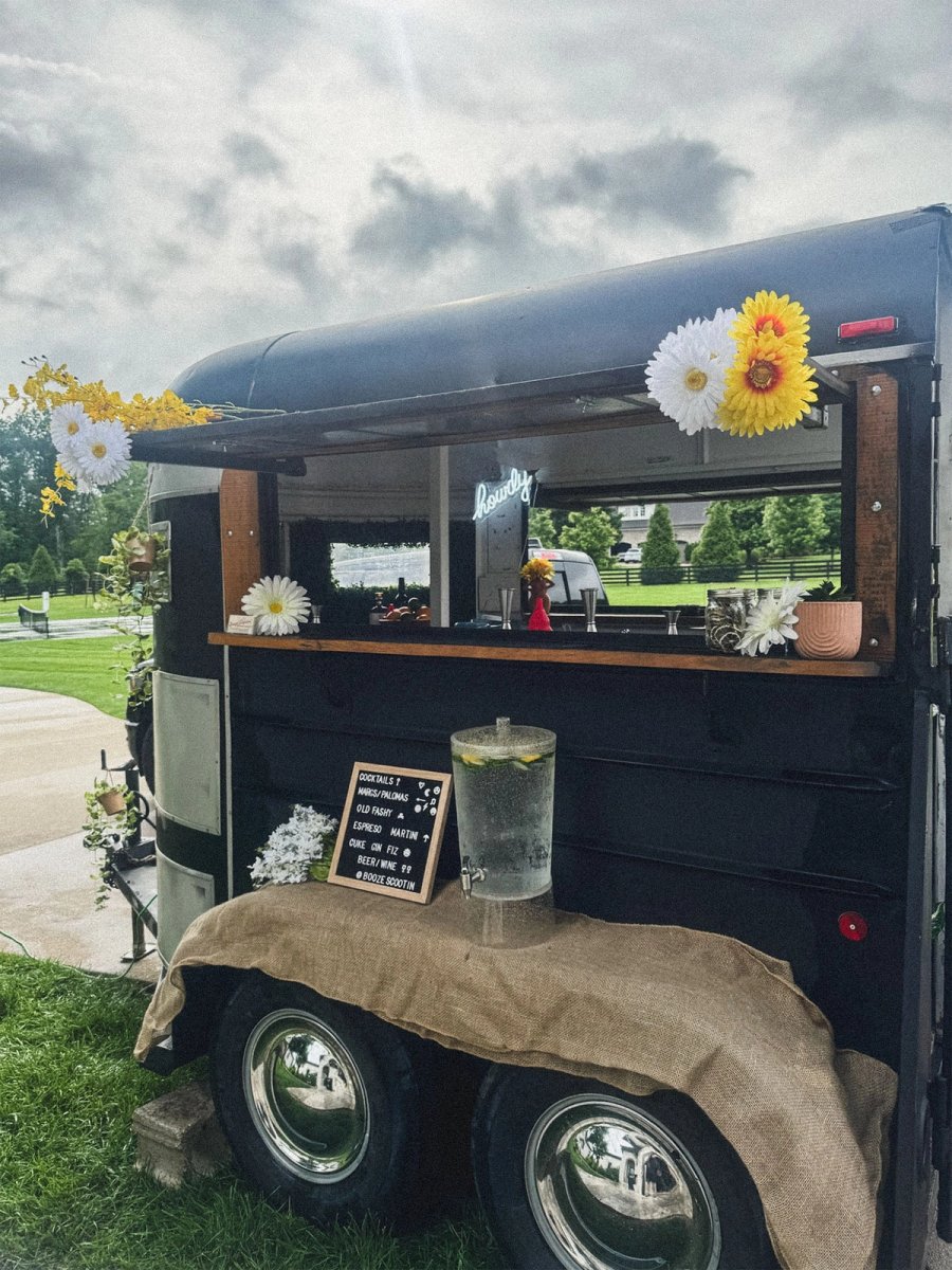 The Howdy trailer parked in a field, daisies along the roof and window, drink dispenser and chalkboard menu out front.