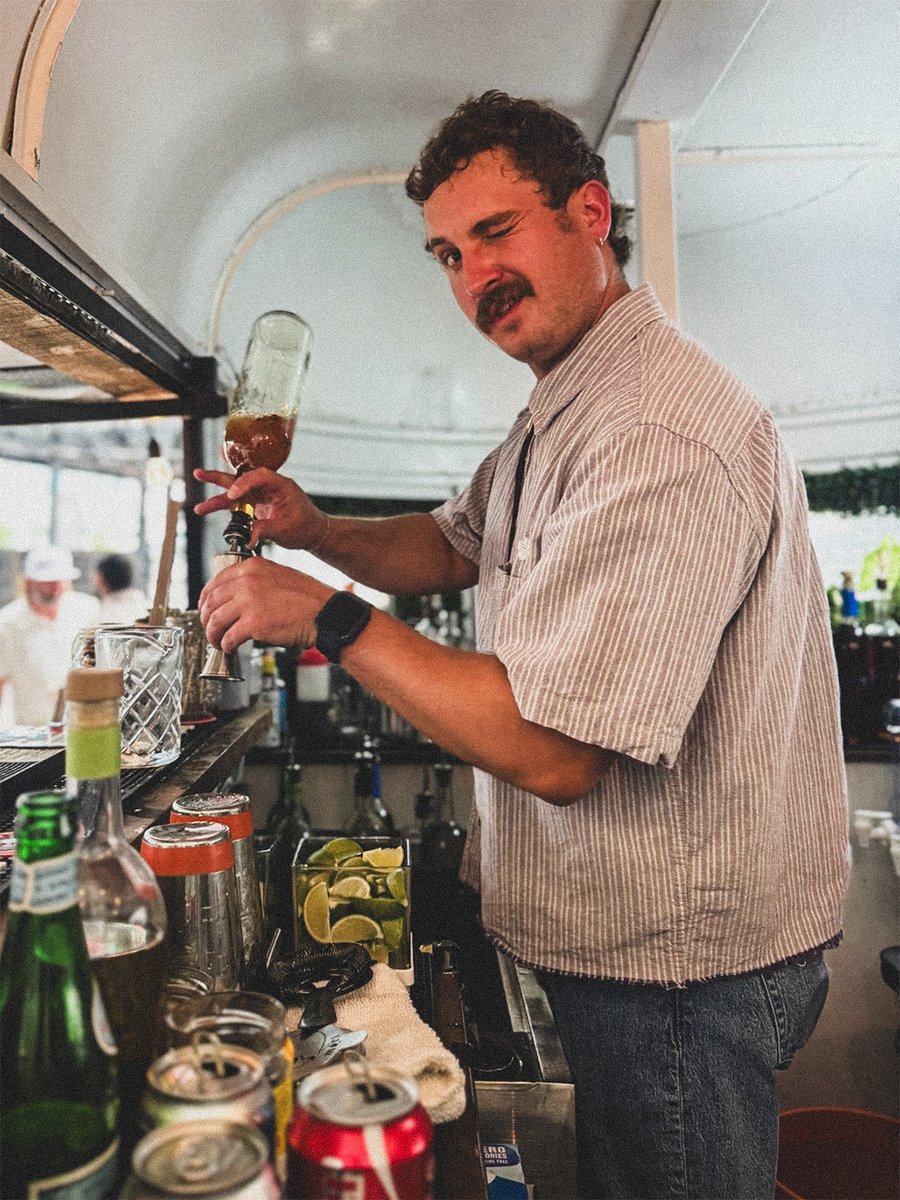 A mustached bartender in a striped shirt tips a beer glass under the tap, bottles and lime slices on the bar.